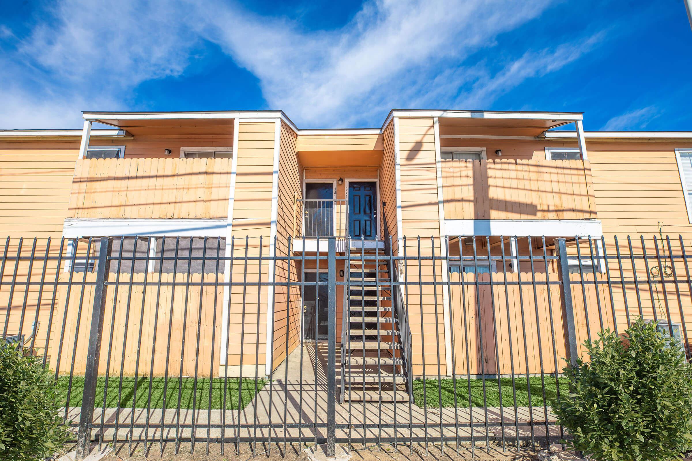 Two-story apartment building with wooden exterior and a blue front door. The entrance is flanked by stairs leading up to a second-floor balcony. The building is surrounded by a black wrought iron fence and features green grass in the front area. The sky is clear with a few clouds.