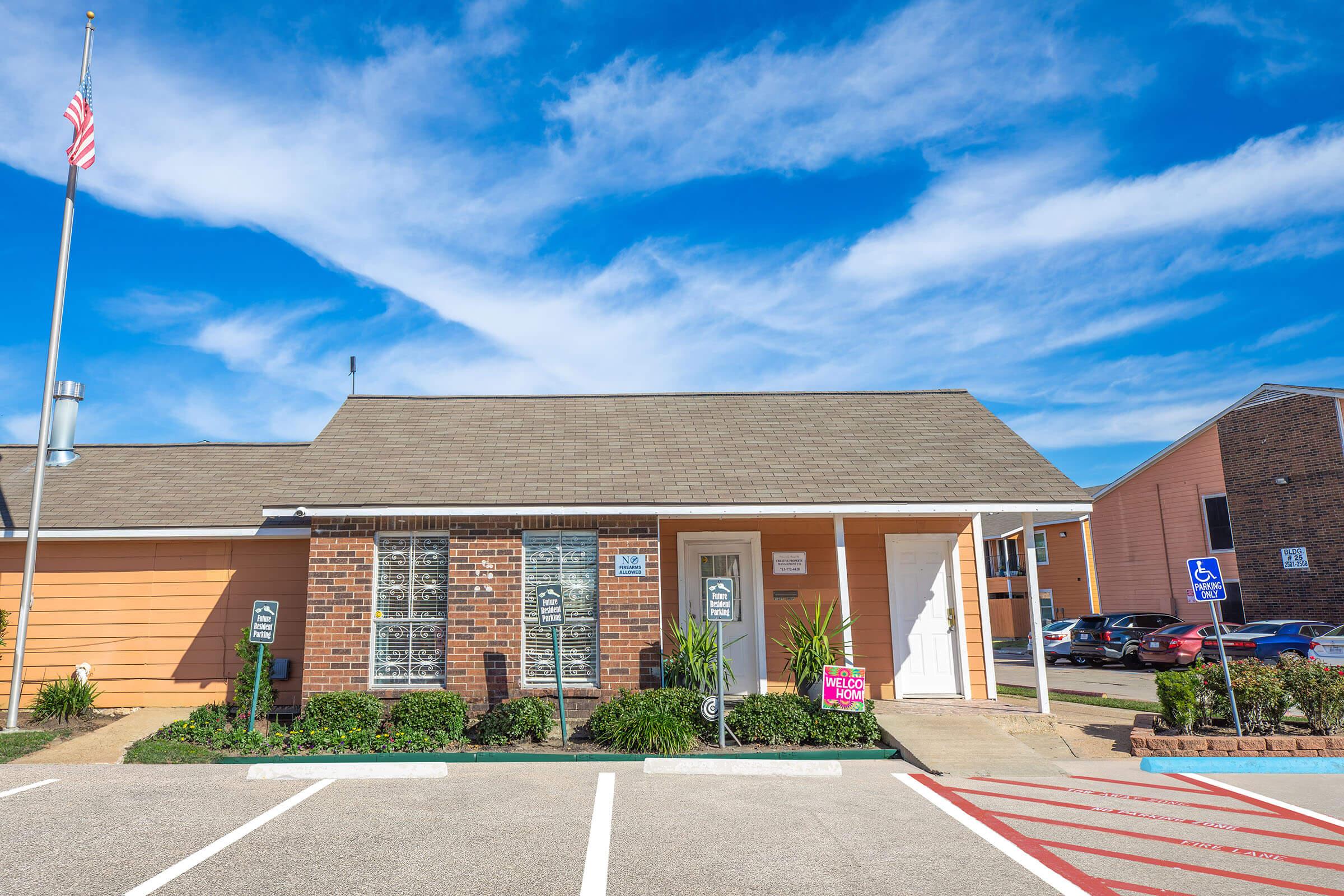 A single-story building with a mix of brick and orange siding, featuring several windows with white shutters. A flagpole stands in front, displaying the American flag. The parking lot has designated spaces, including accessible parking signs. Lush green landscaping lines the entrance, and a sign welcomes visitors.