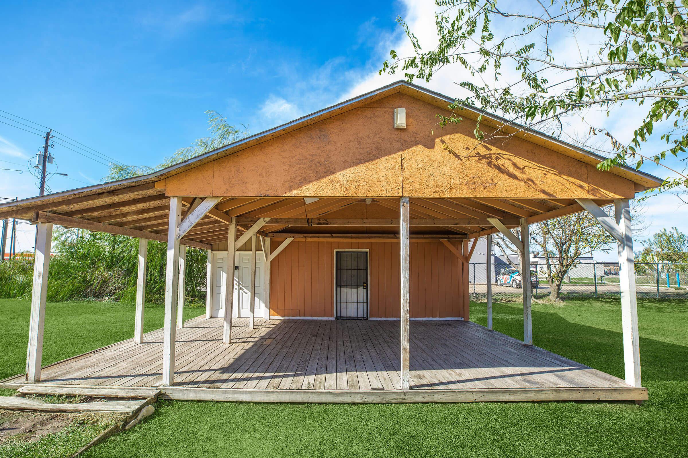 A wooden pavilion with an orange exterior and a slanted roof, featuring open sides and a wooden deck. The surrounding area has green grass and a clear blue sky. There are a few trees and utility poles in the background, suggesting a rural or park-like setting.