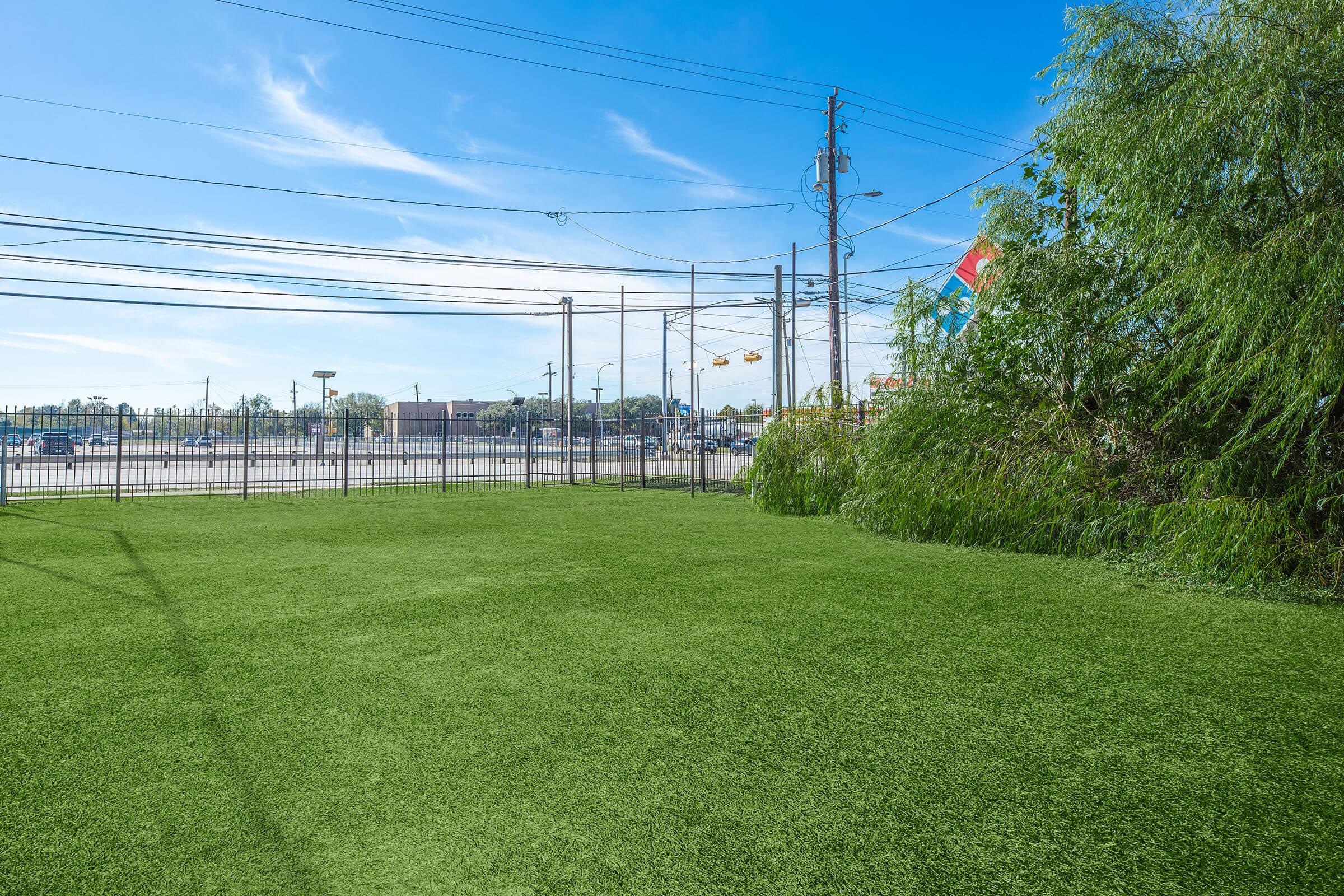 A wide view of a green artificial grass area with a clear blue sky overhead. In the background, there are utility poles and power lines, along with a fenced boundary. Sparse trees and a hint of a parking lot can be seen in the distance. The scene appears open and well-maintained.