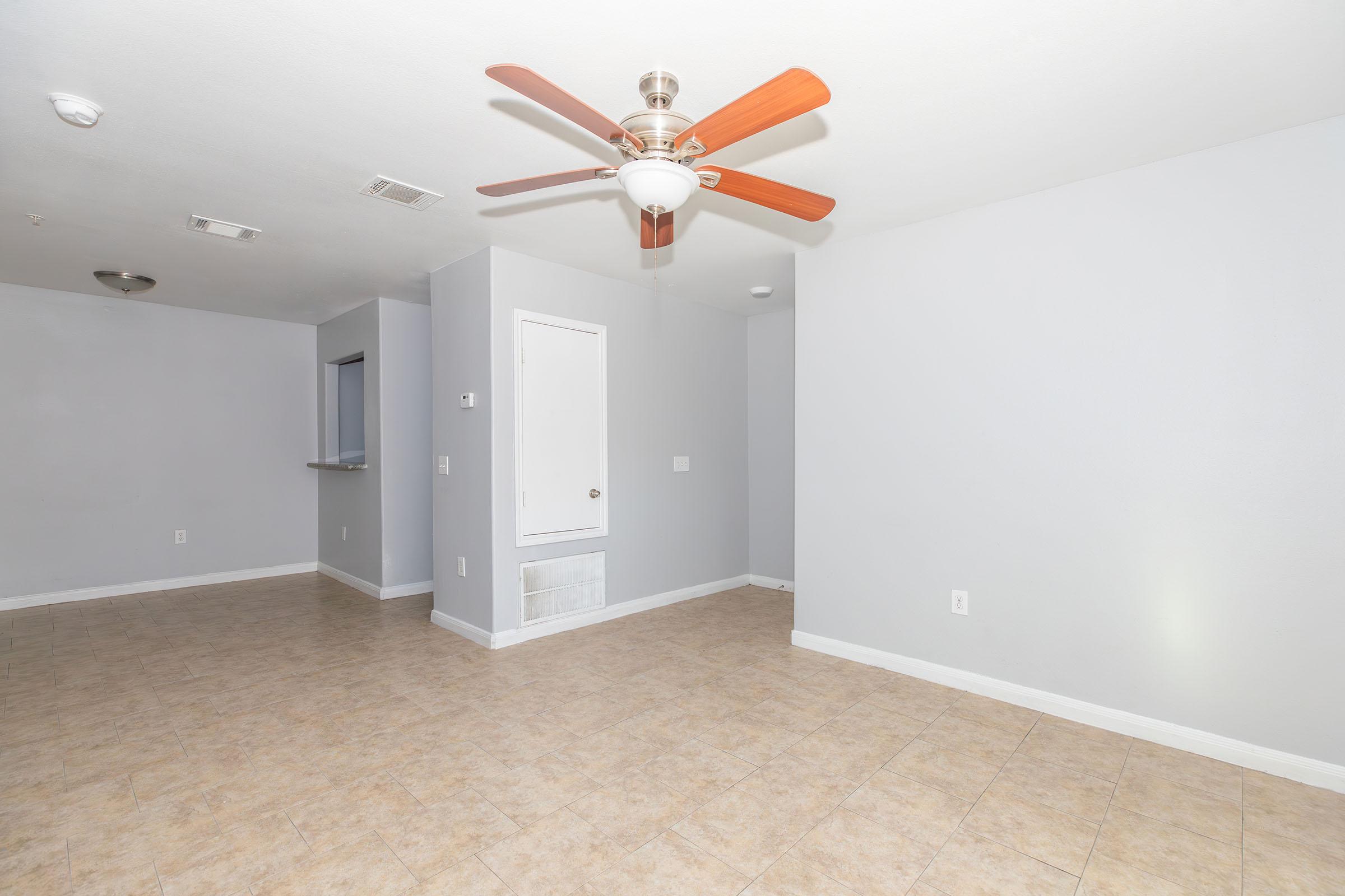 A clean, empty room featuring light gray walls and tile flooring. It has a ceiling fan with wooden blades, a closed door leading to another area, and a small wall cutout. The space is well-lit and appears to be designed for a living or dining area.