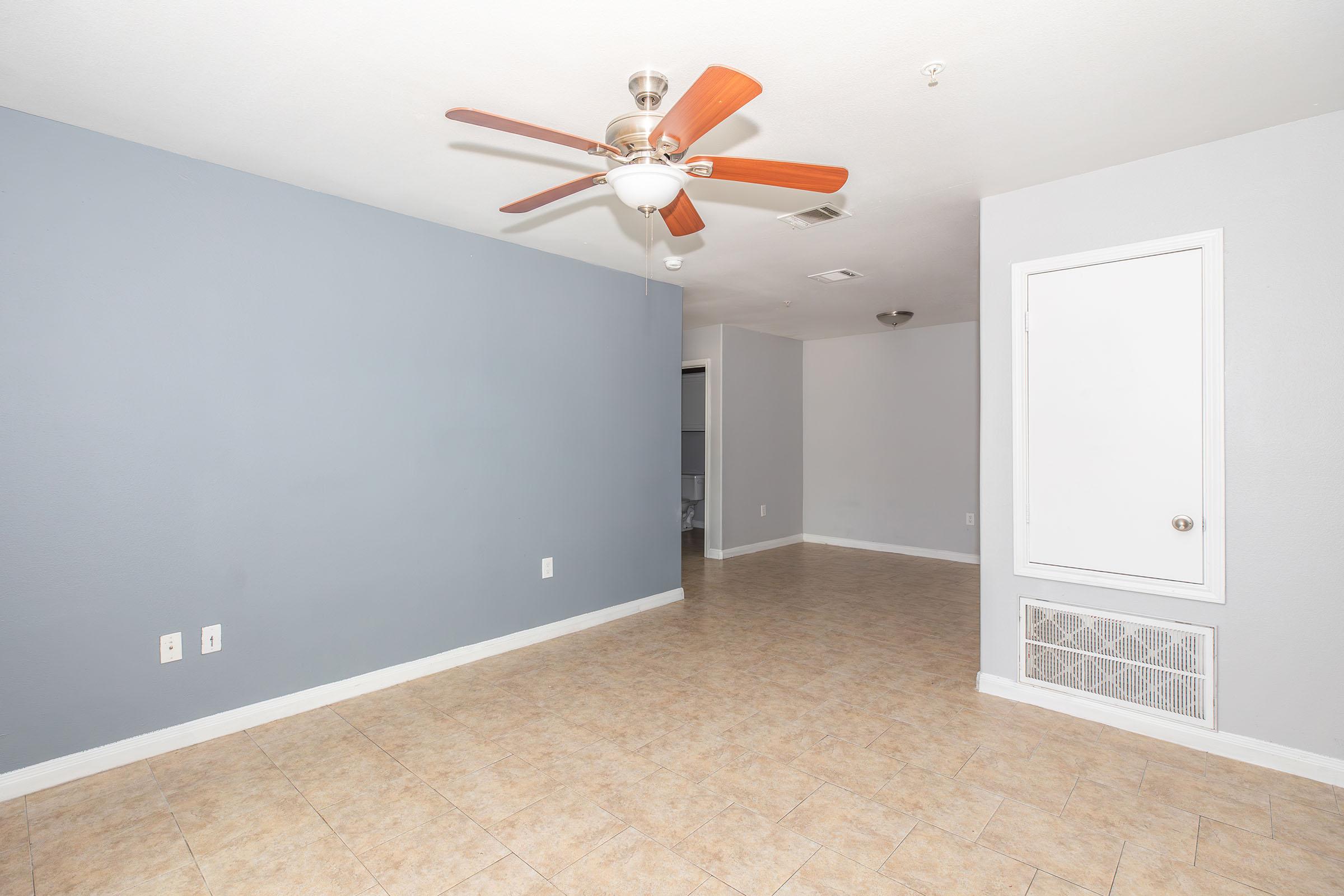 Interior view of a spacious room with light gray walls, a ceiling fan with orange blades, and tiled flooring. There is a white air vent on the floor and a closed white door in the corner. The area is well-lit and features a minimalistic design, suitable for various layouts.