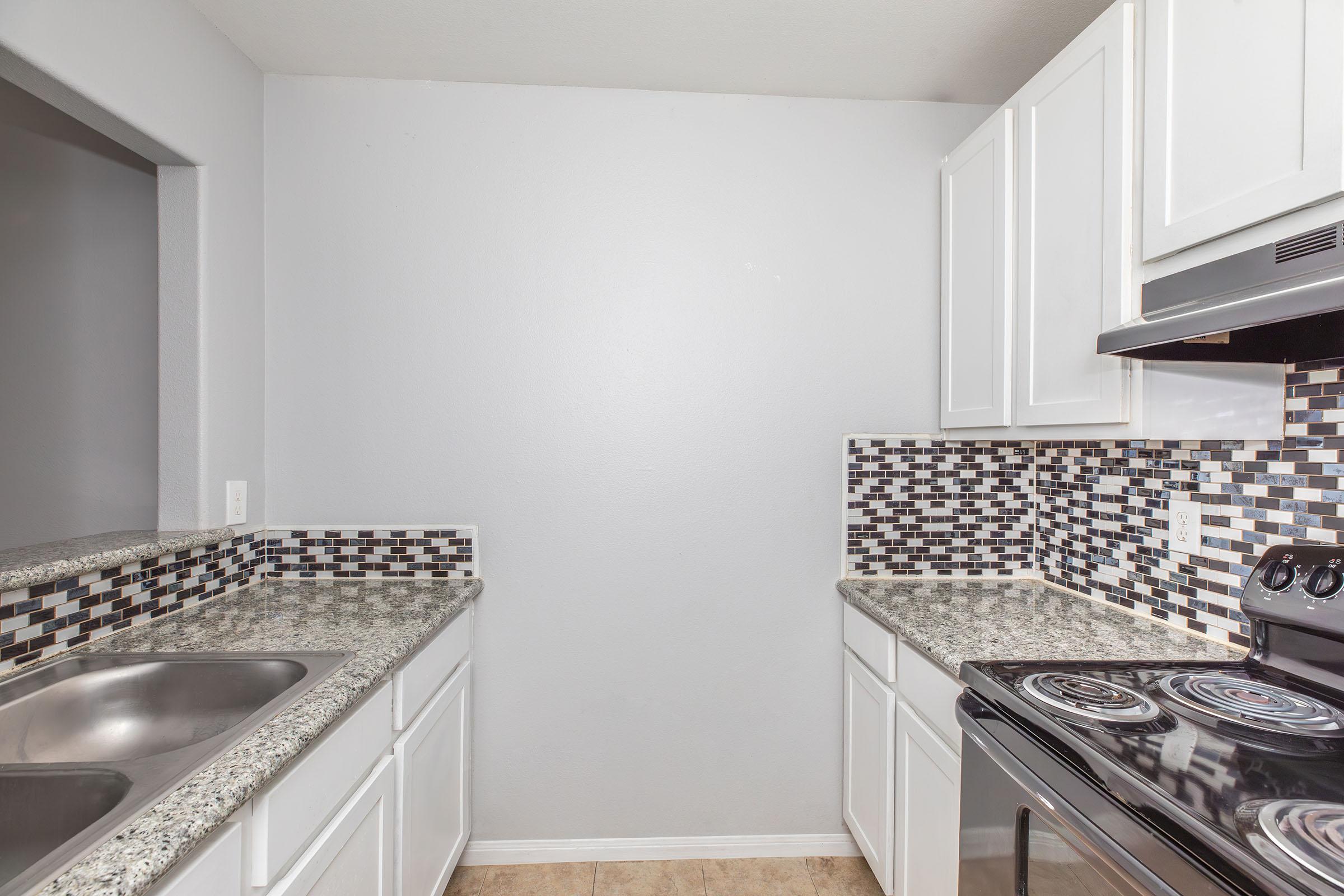 A modern kitchen featuring white cabinetry, a black stove, and a granite countertop. The backsplash consists of a mosaic pattern in shades of gray and black. The kitchen is well-lit with a neutral wall color and has an open layout, showcasing a clean and organized space.