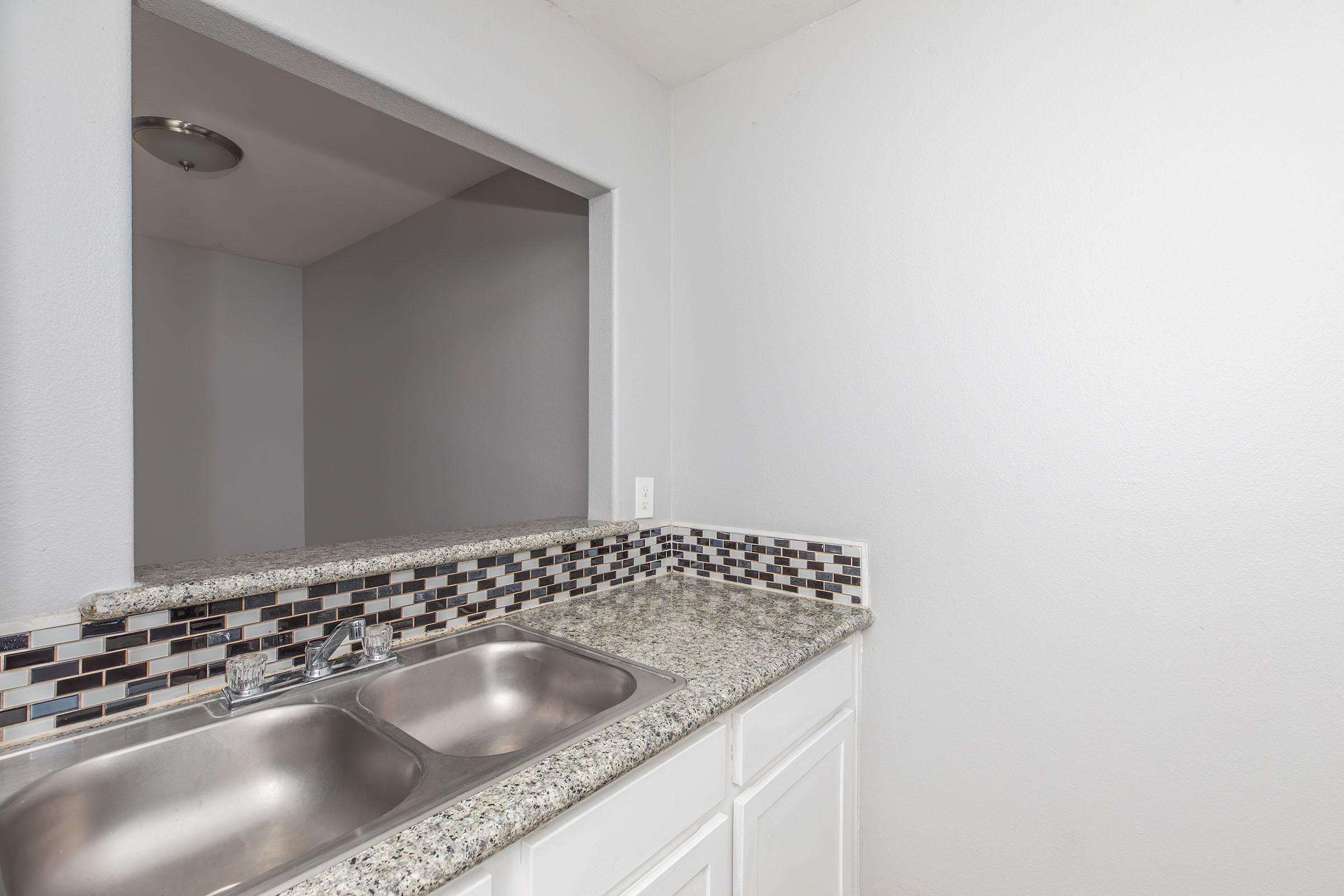 Kitchen area featuring a double stainless steel sink set into a granite countertop. The backsplash consists of black, white, and gray tiles. The walls are painted a light gray, and there is an overhead light fixture visible above the sink area. The space appears clean and modern.