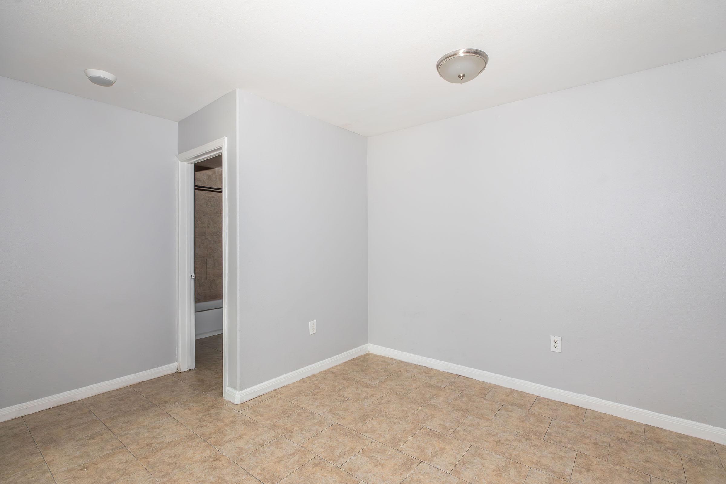 Empty room with light gray walls and tiled flooring. A single light fixture is mounted on the ceiling. There is a door on the left leading to another space, and no furniture or decorations are visible, creating a spacious and minimalist appearance.