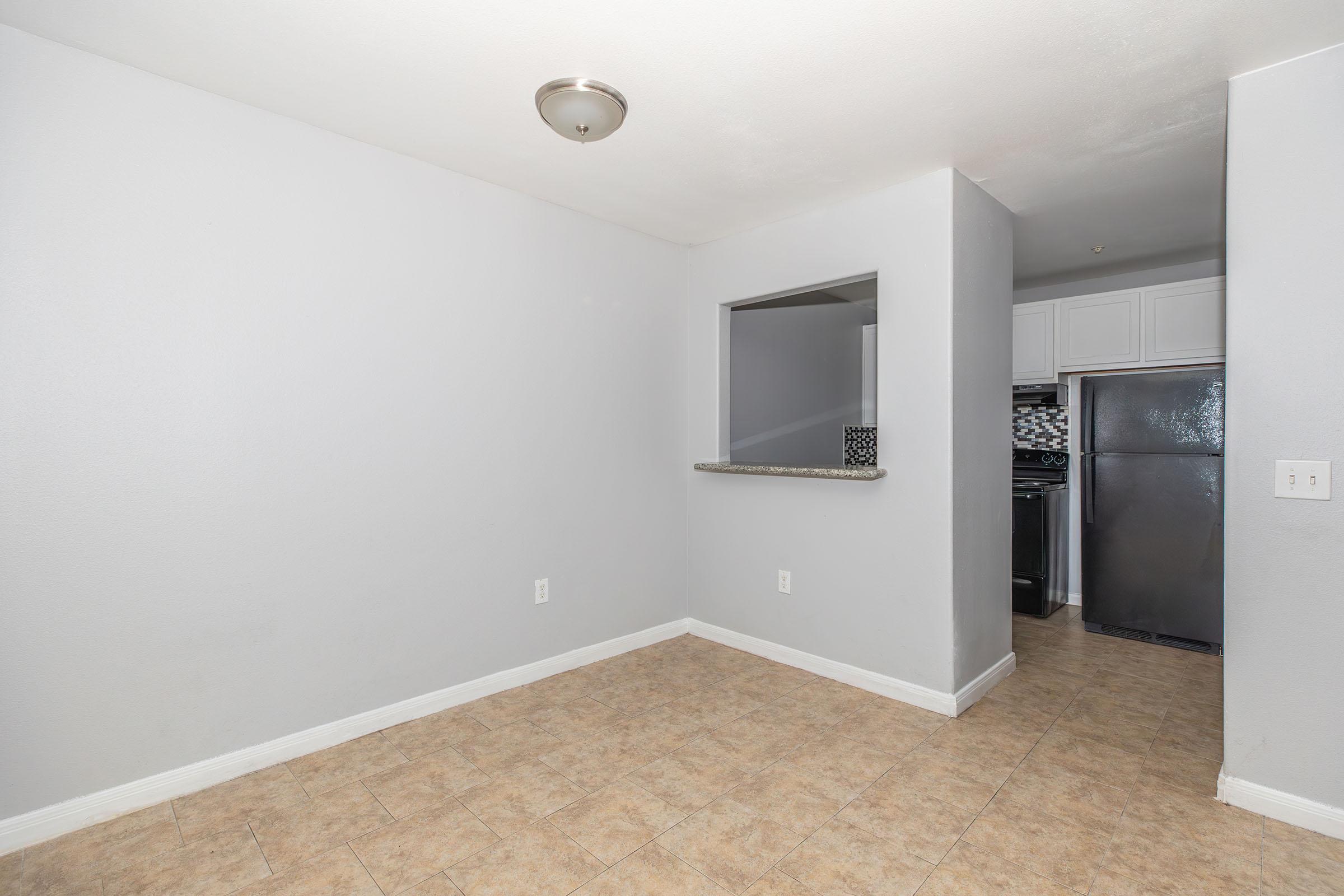 Empty room with light gray walls and tiled flooring. A small counter separates the room from a kitchen area visible in the background, featuring dark appliances. The lighting is soft, and there are no furnishings or decorations present, creating a spacious and minimalist atmosphere.