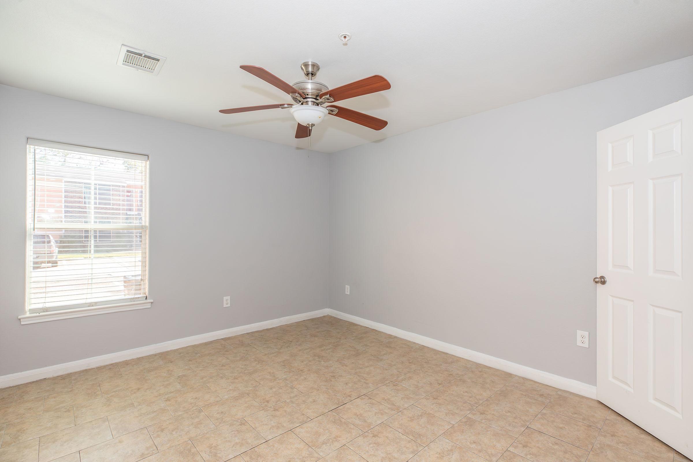 Empty room featuring light gray walls, a ceiling fan with wooden blades, and a window with white blinds. The flooring is a light-colored tile, and there's a closed white door on one side. The space has a bright and airy feel, suitable for various uses.