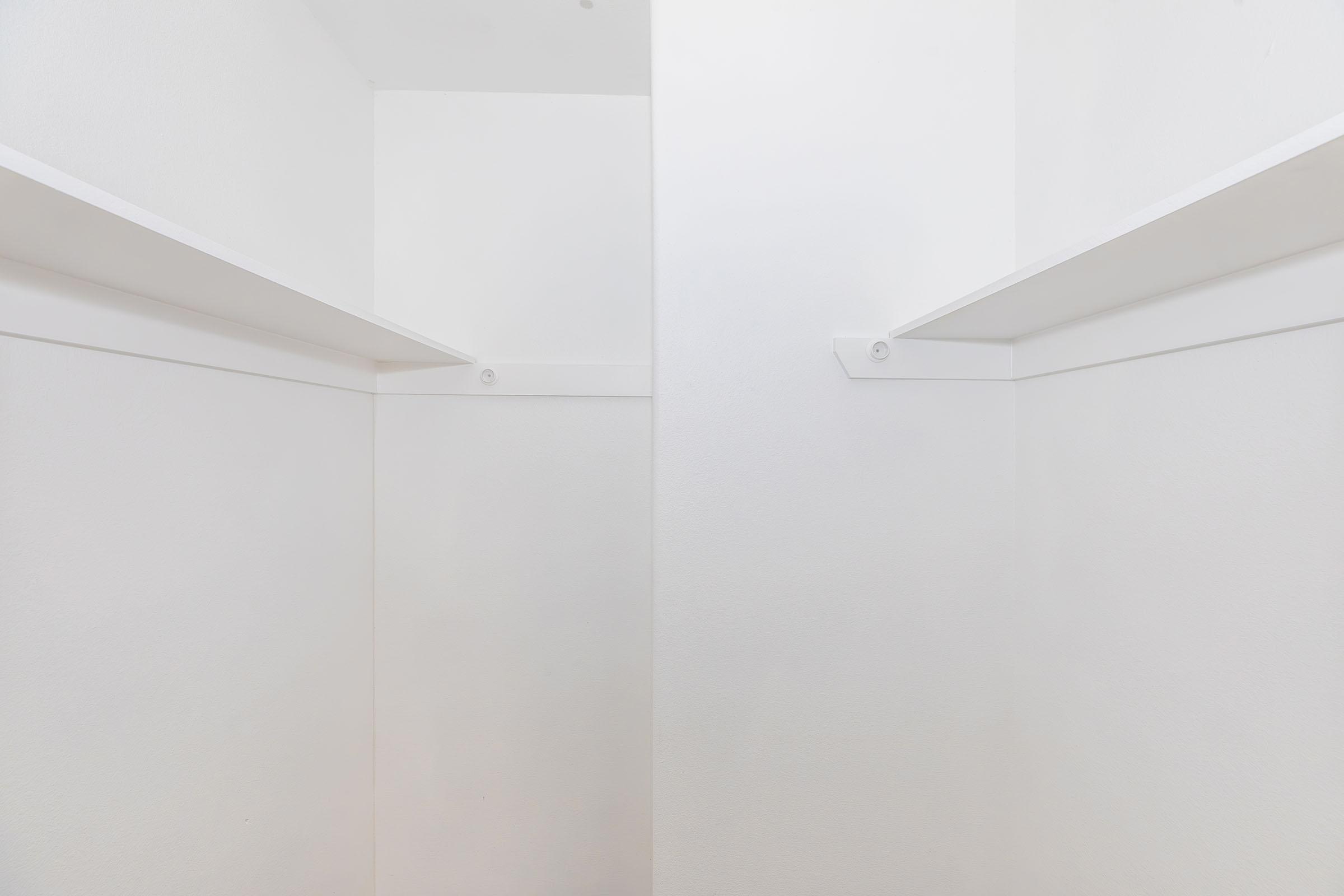 Empty white closet with two walls and shelves, featuring a minimalist design and bright lighting. The shelves are mounted at equal heights on either side, providing a clean and airy appearance. The overall color scheme is entirely white, contributing to a sense of spaciousness.