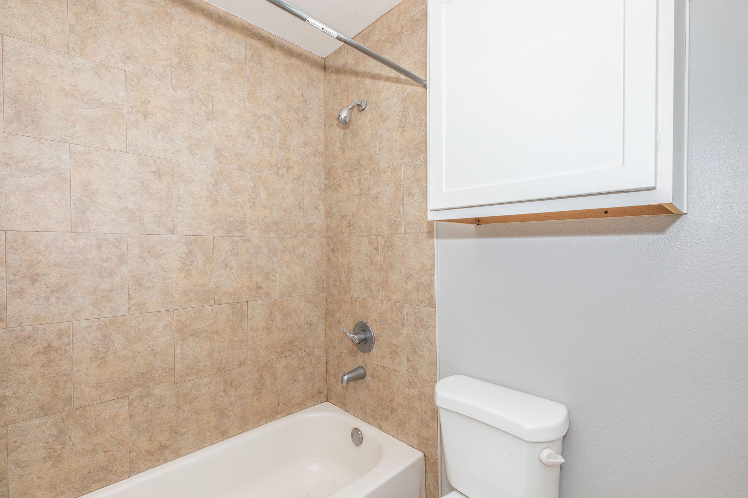 A clean bathroom featuring a beige and brown tiled wall, a bathtub with a showerhead above, a wall-mounted cabinet, and a white toilet. The space is well-lit and neatly arranged, showcasing a simple and functional design.