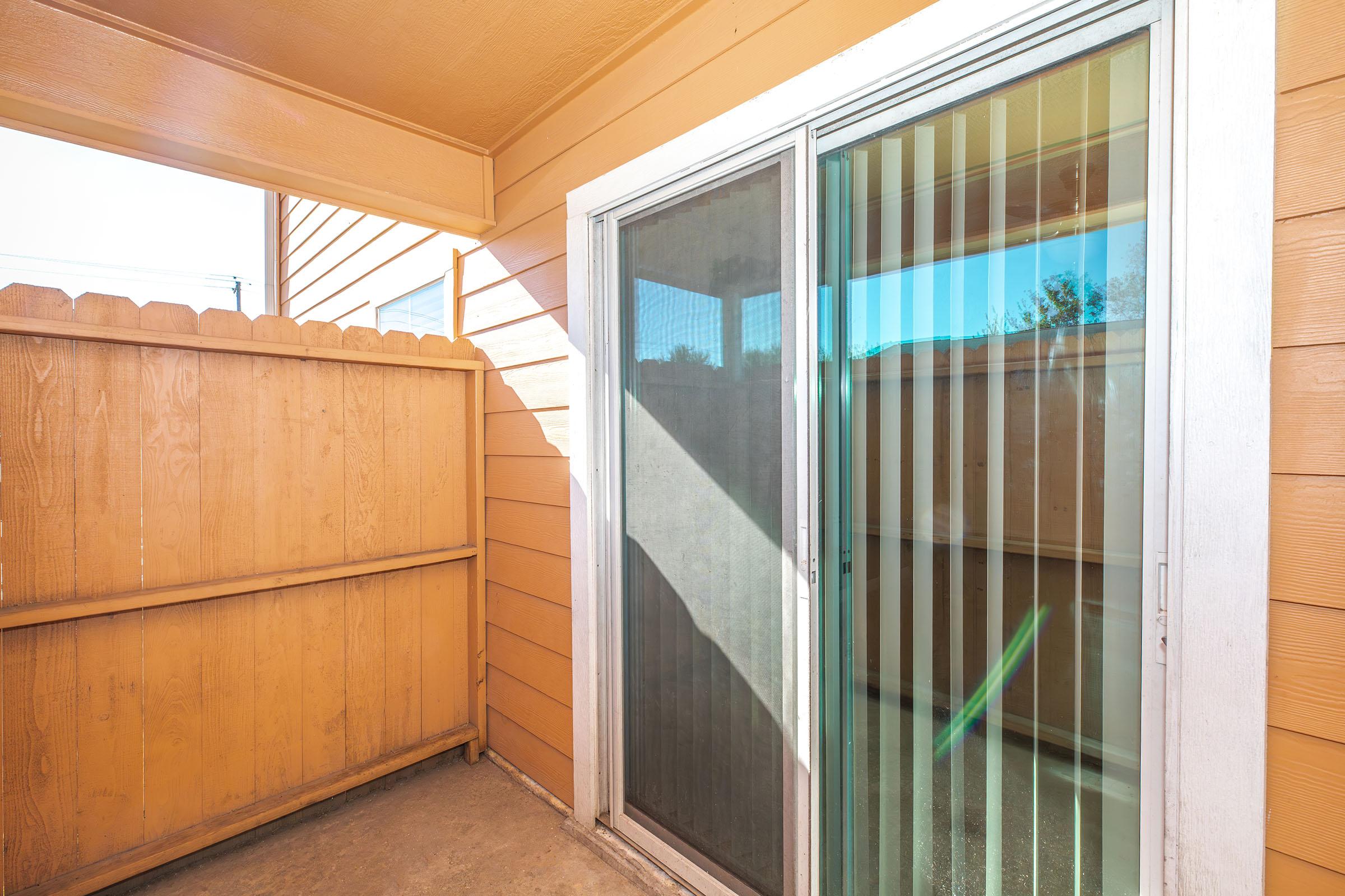 A view of a small outdoor balcony or patio area with wooden fencing, featuring sliding glass doors with vertical blinds. The walls are painted a warm orange color, and natural light illuminates the space, creating a bright and inviting atmosphere.