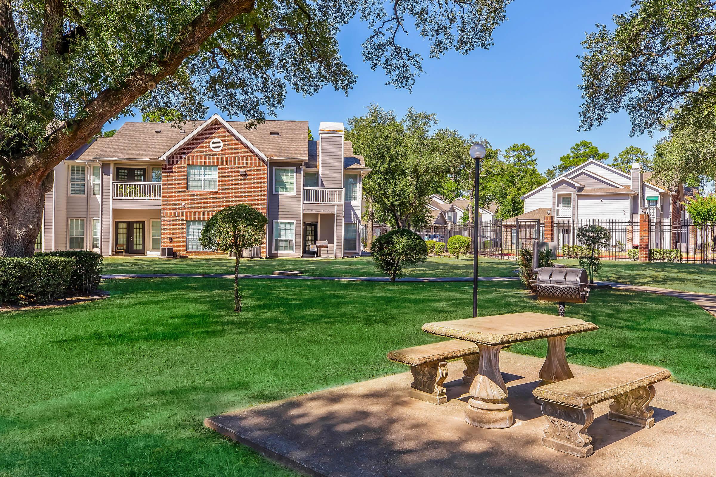 A serene outdoor area featuring a stone picnic table surrounded by lush green grass, small hedges, and trees. In the background, there are apartment buildings with balconies and a well-maintained pathway leading to a lamppost, creating an inviting atmosphere for relaxation.