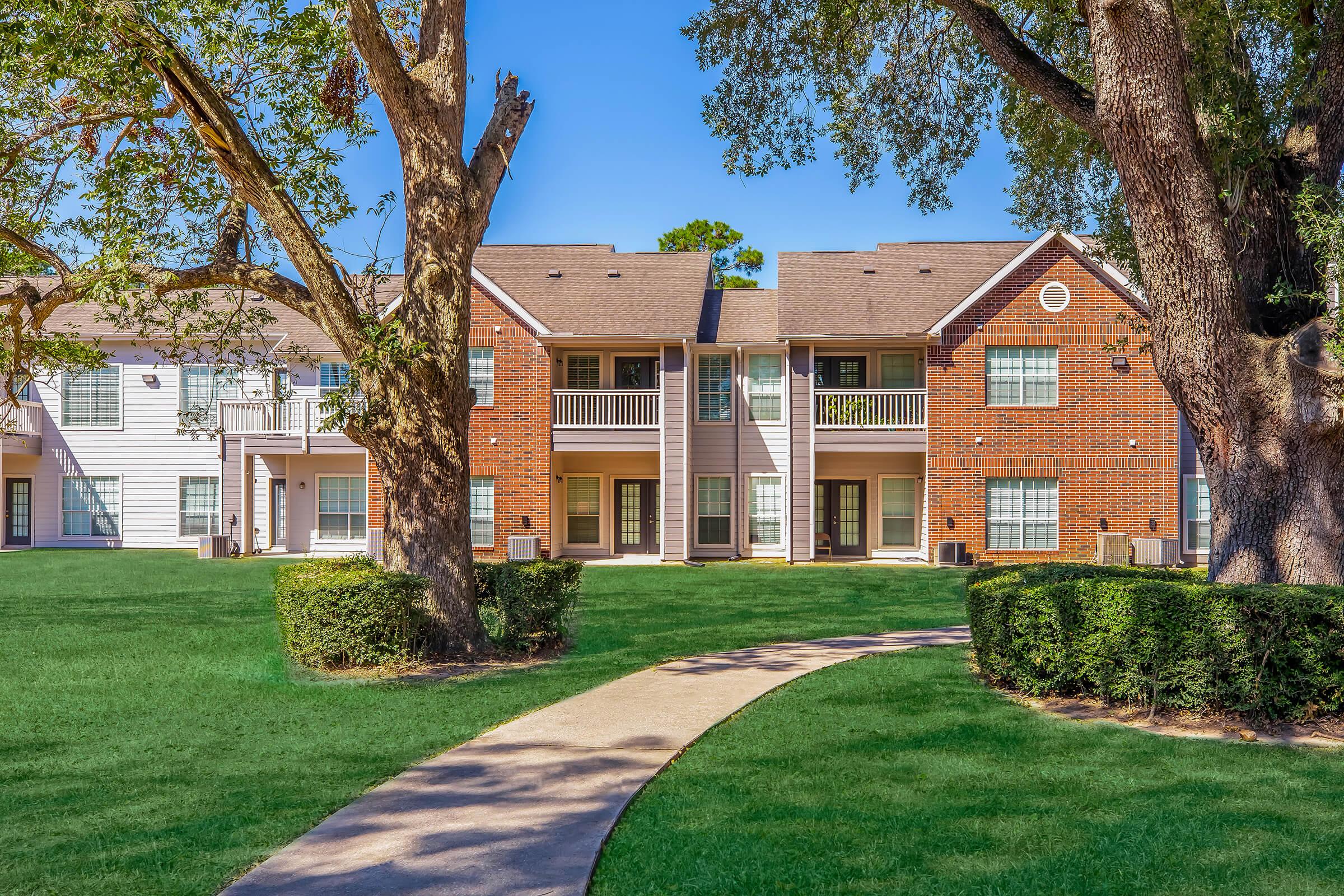 A well-maintained apartment complex surrounded by lush green grass and trees, featuring two buildings with a mix of brick and white siding. A paved pathway leads through the grassy area, with balconies visible on the upper floors of the buildings under a clear blue sky.