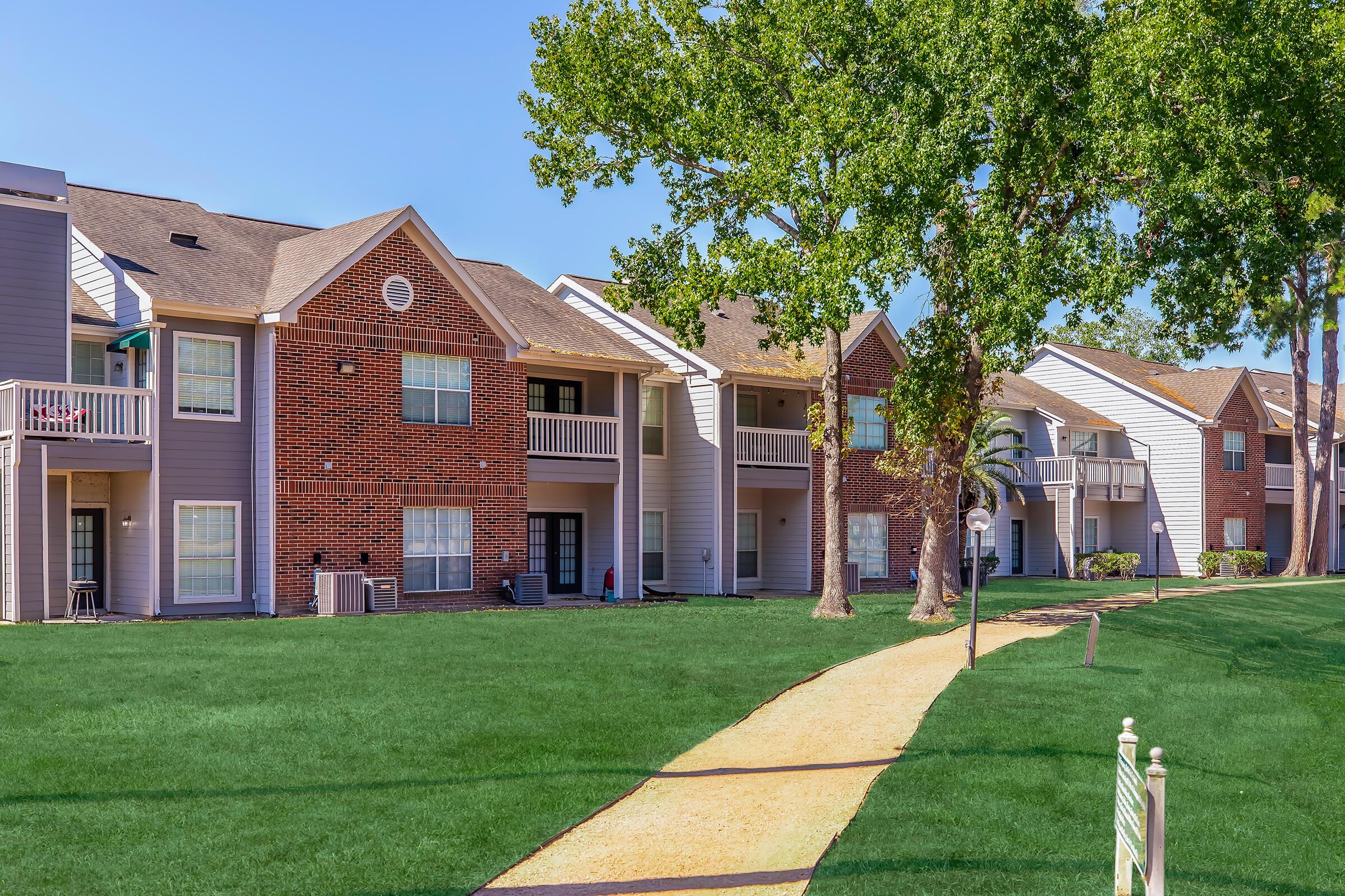 A well-maintained apartment complex featuring multiple buildings with a mix of brick and siding exteriors. Green lawns surround the buildings, and a curved gravel path leads through the landscape. Trees provide shade, and some balconies are visible on the upper floors. The sky is clear and blue.