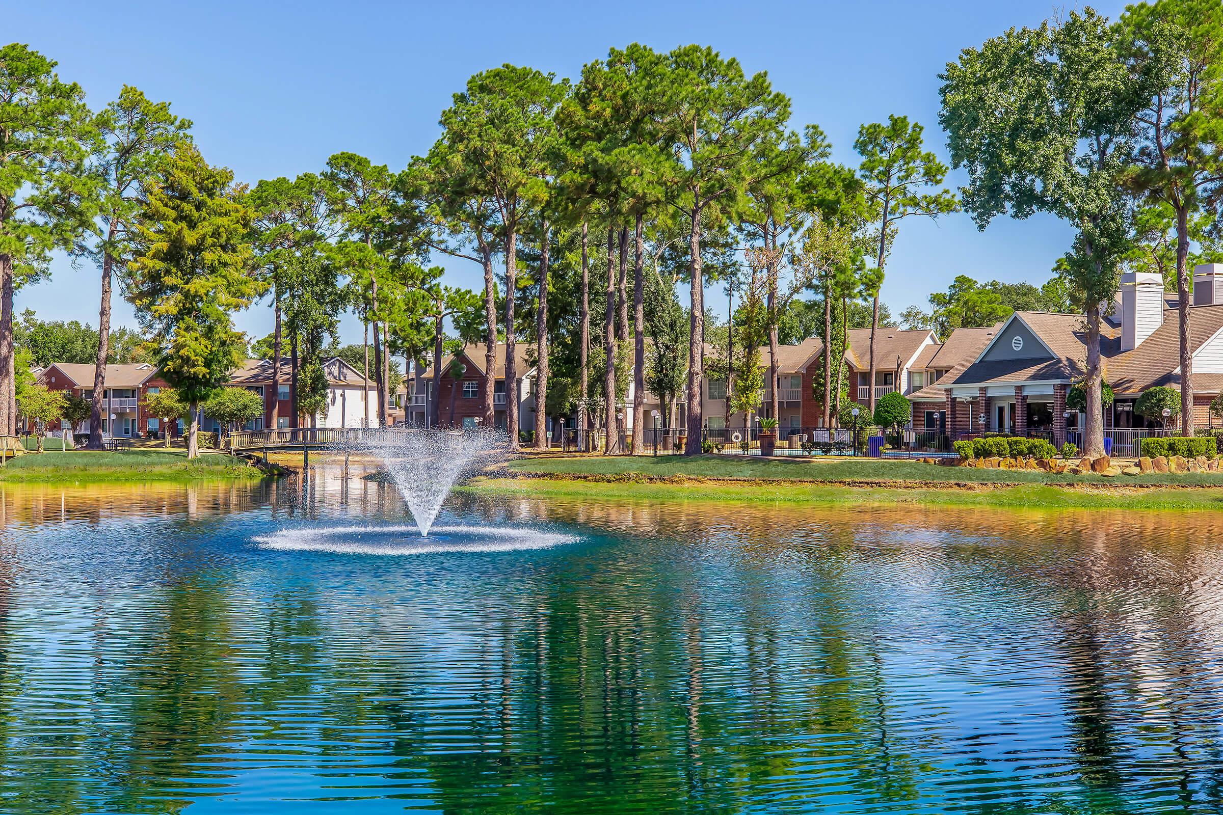 A serene lake surrounded by lush green trees and residential buildings. In the center, a fountain sprays water into the air, creating a tranquil atmosphere. The clear blue sky reflects on the water's surface, enhancing the peaceful setting.