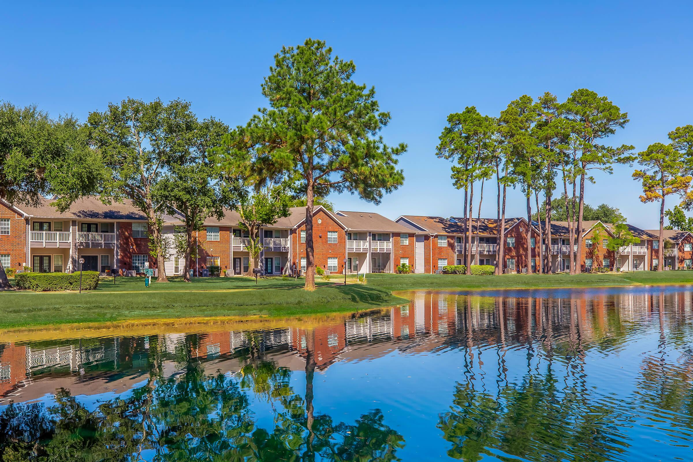 A serene lakeside view of a residential apartment complex with brick buildings surrounded by lush green trees. The clear blue sky reflects in the calm water, creating a tranquil atmosphere. The scene showcases well-maintained lawns and a peaceful environment.