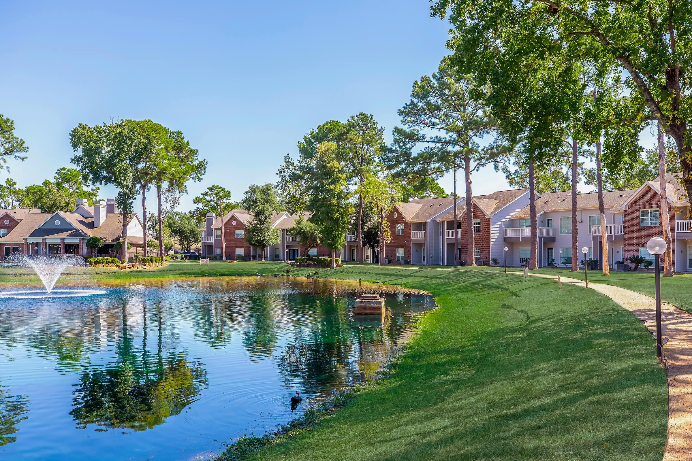 A serene landscape featuring a pond surrounded by lush greenery and residential buildings. A fountain sprays water in the center of the pond, with trees lining the shore and a clear blue sky above. The path along the water invites leisurely walks in this tranquil setting.
