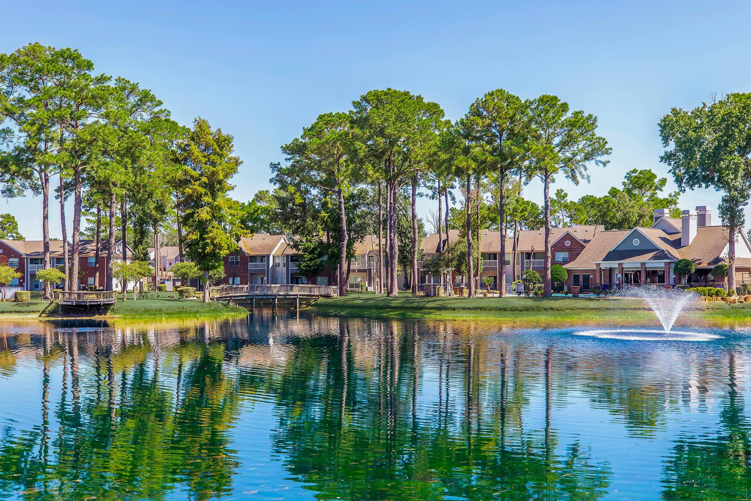 A serene lake surrounded by tall pine trees and residential buildings. A fountain creates ripples in the water, reflecting the clear blue sky and the greenery. The scene conveys tranquility and natural beauty in a suburban setting.
