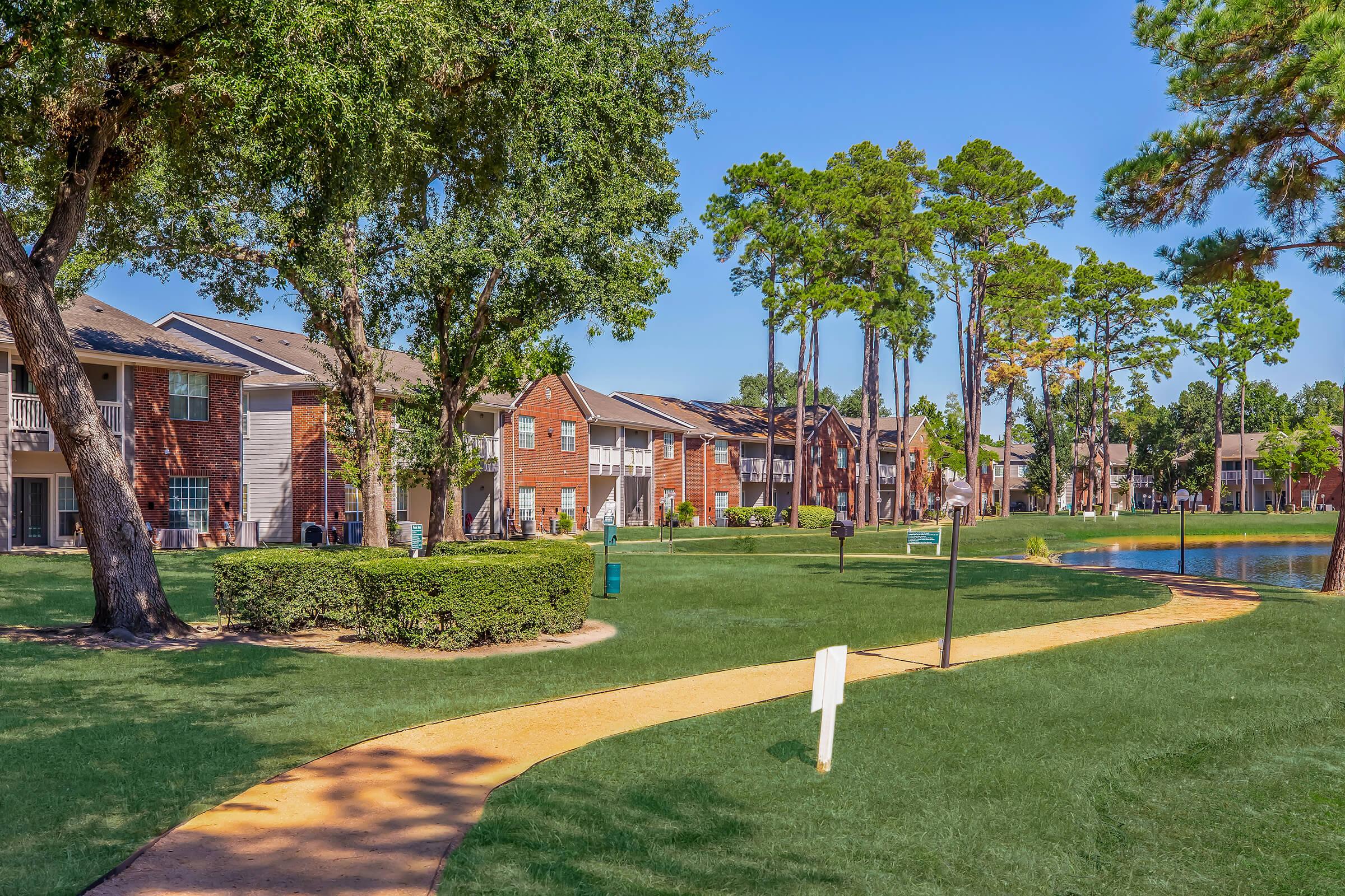 Lush green lawn with a winding path leading through a residential apartment complex. The scene features brick buildings surrounded by tall trees, and a calm pond in the background, under a clear blue sky. The inviting landscape suggests a peaceful suburban environment.