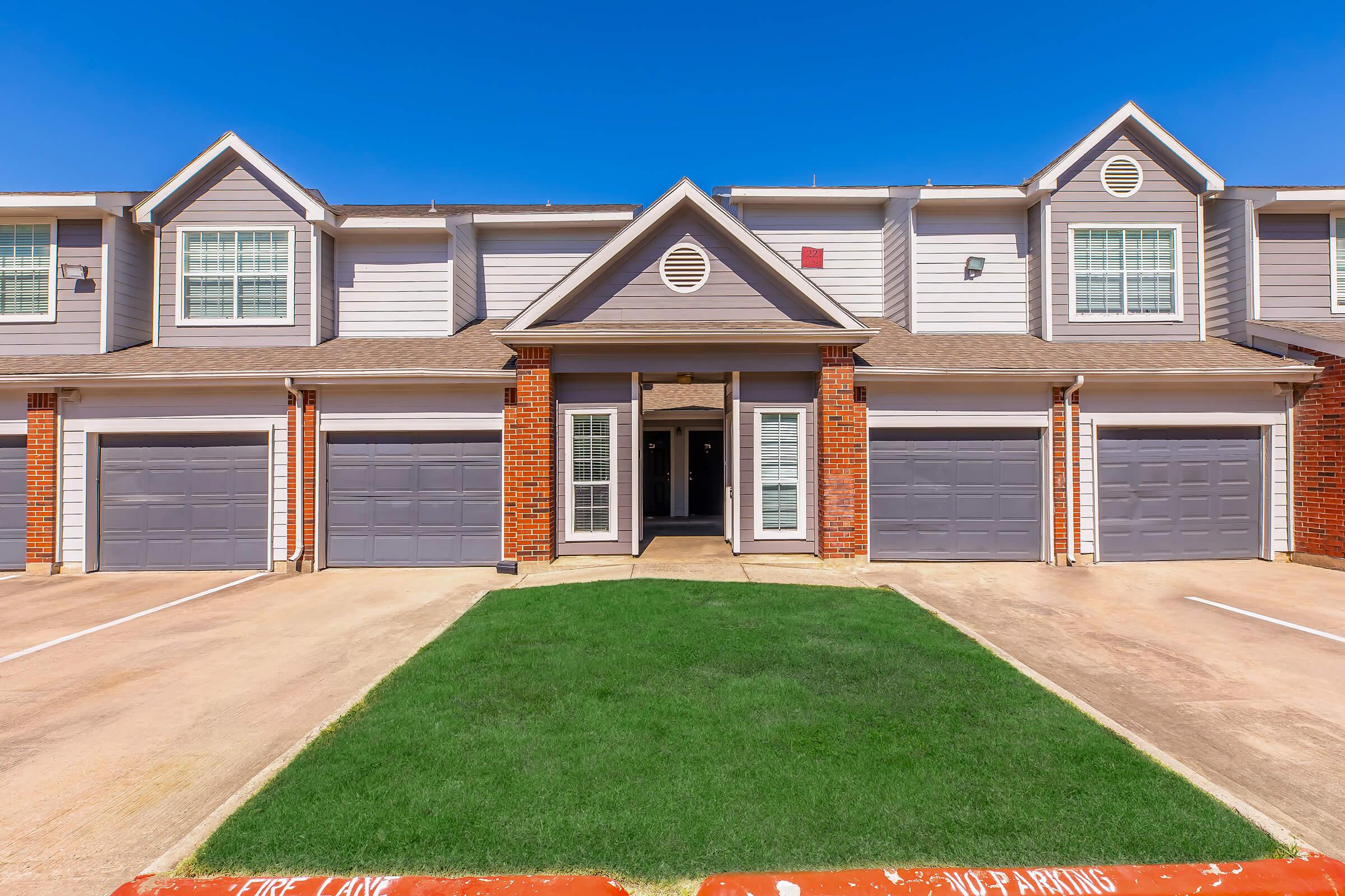 A modern townhouse with gray siding and brick accents. It features multiple garages, a well-maintained front lawn, and a clear blue sky. The entrance has double doors flanked by large windows, creating an inviting appearance. Parking spaces are visible in front of the units.