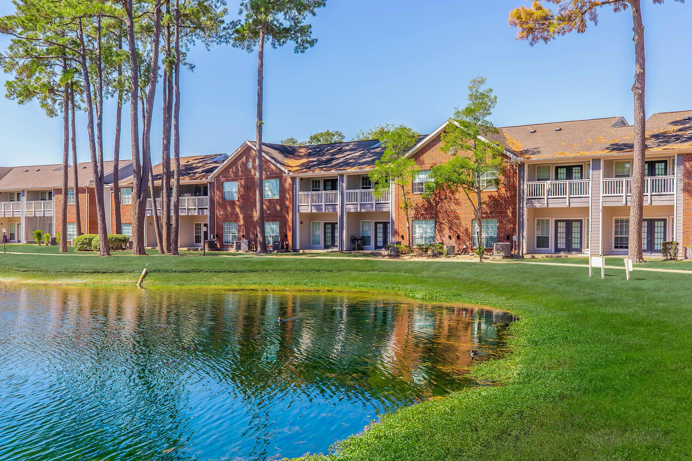 A picturesque view of well-maintained apartment buildings beside a serene pond, surrounded by lush green grass and tall trees. The clear blue sky reflects on the water's surface, enhancing the tranquil atmosphere of the residential area.