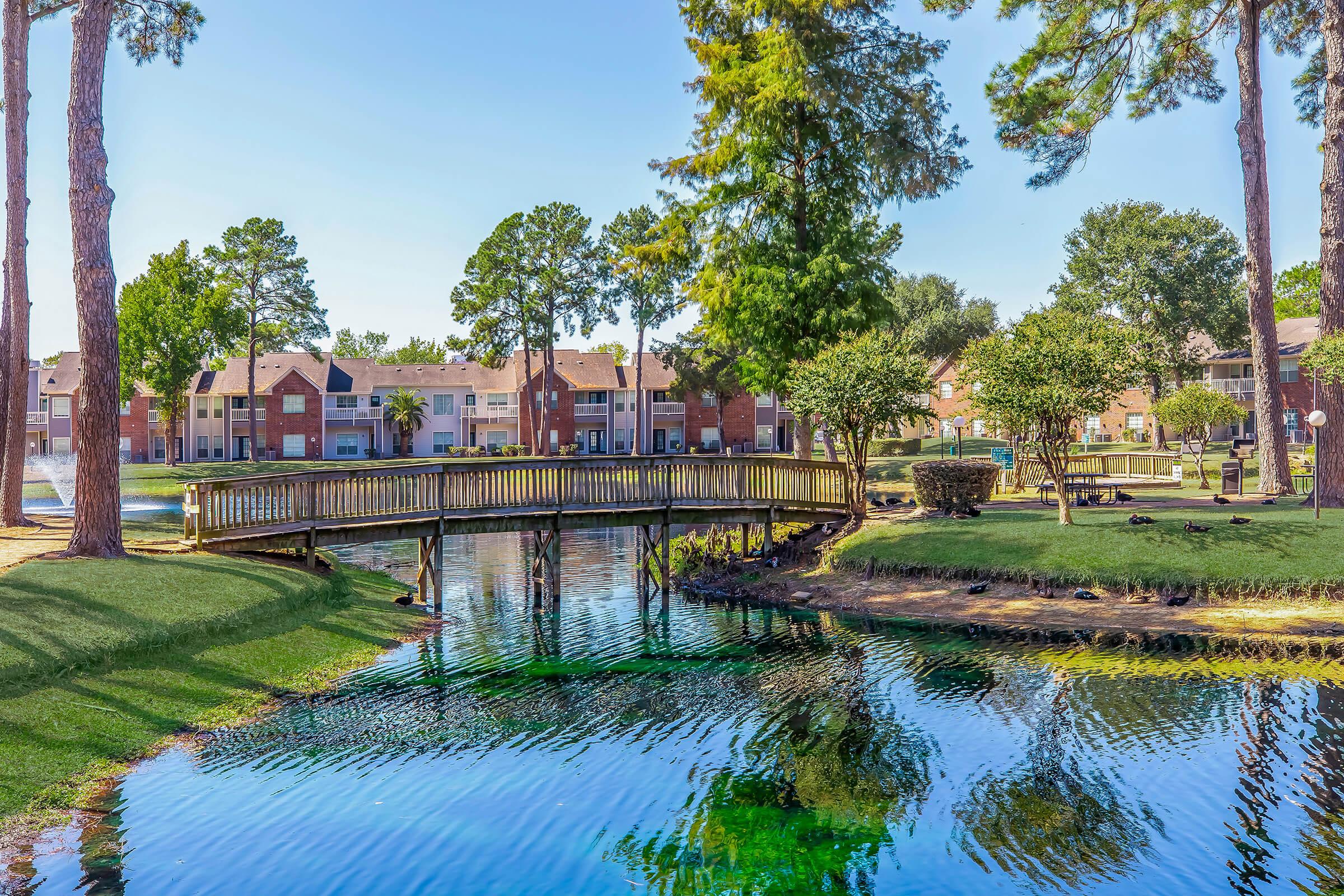 A serene landscape featuring a wooden footbridge over a clear, reflective pond. Surrounding the pond are lush green lawns and trees, with apartment buildings visible in the background. The scene is peaceful, highlighting the natural beauty of the area.