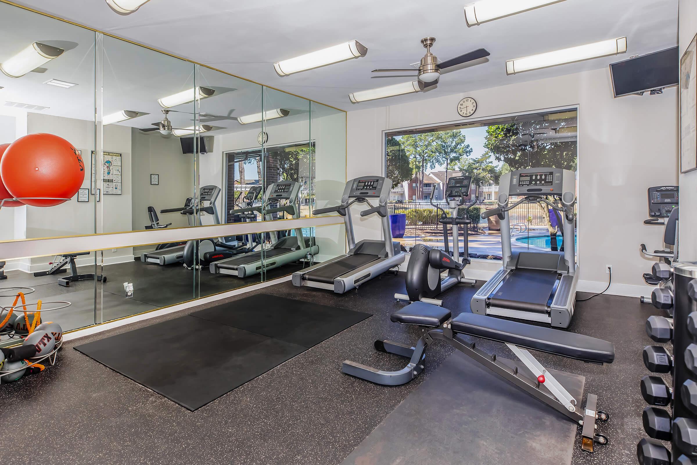 A modern gym interior featuring two treadmills facing large windows, a weight bench, and exercise equipment. Mirrors line one wall, and a large red exercise ball is positioned nearby. Ceiling fans and a clock add to the functional workout space.