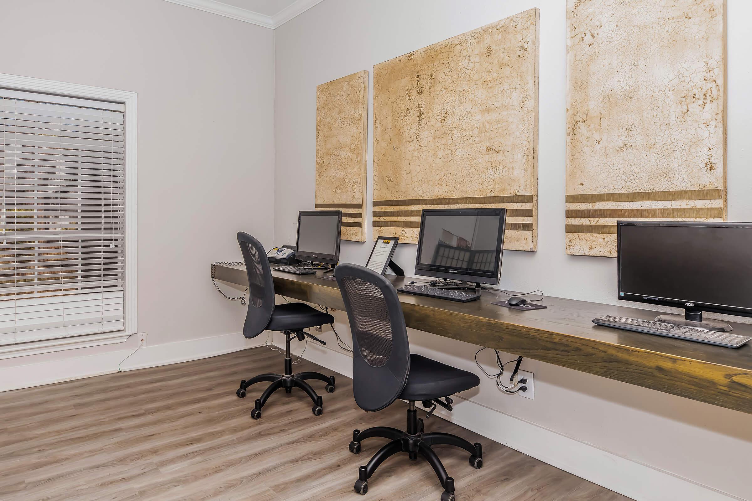 A modern office space featuring two workstations with desktop computers and ergonomic chairs. The walls are adorned with large abstract art pieces, and there is a window with horizontal blinds allowing natural light to enter. The flooring is light-colored wood.