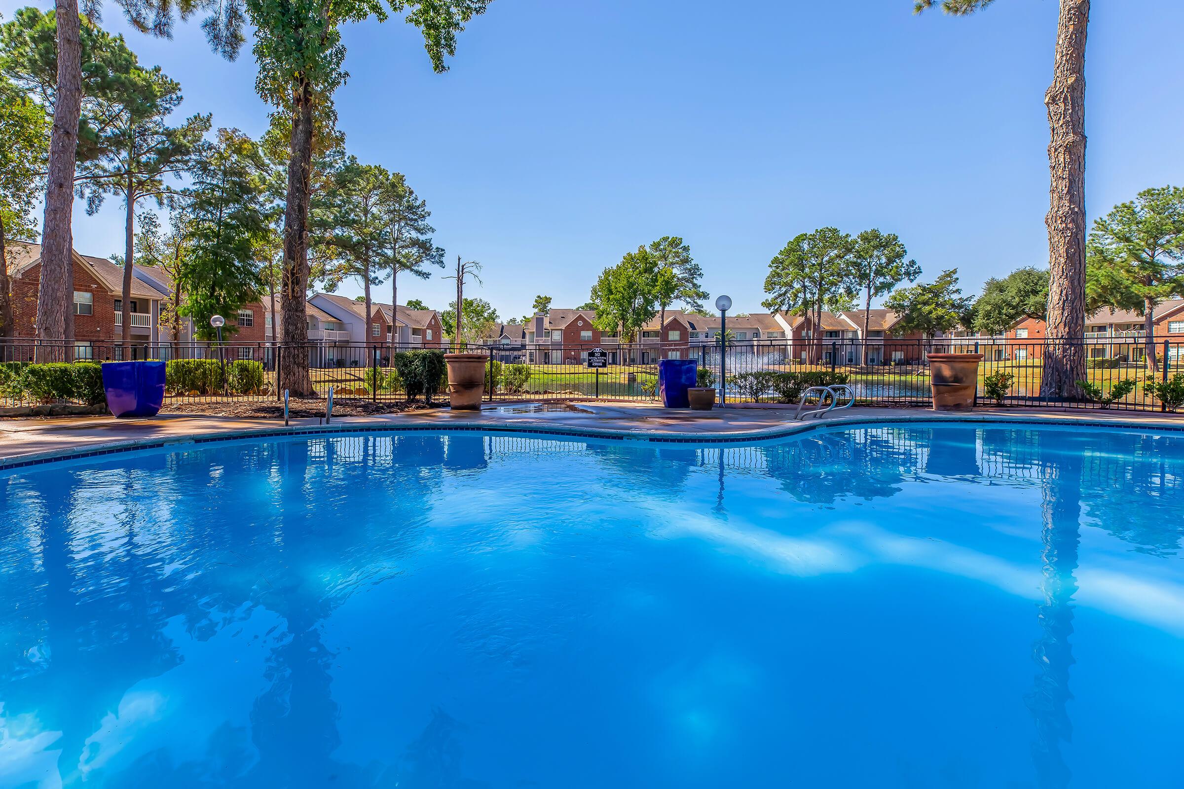 A clear blue swimming pool surrounded by lush greenery and tall trees. In the background, residential buildings and well-maintained lawns are visible, under a bright, sunny sky. The scene conveys a relaxing atmosphere ideal for leisure and outdoor activities.