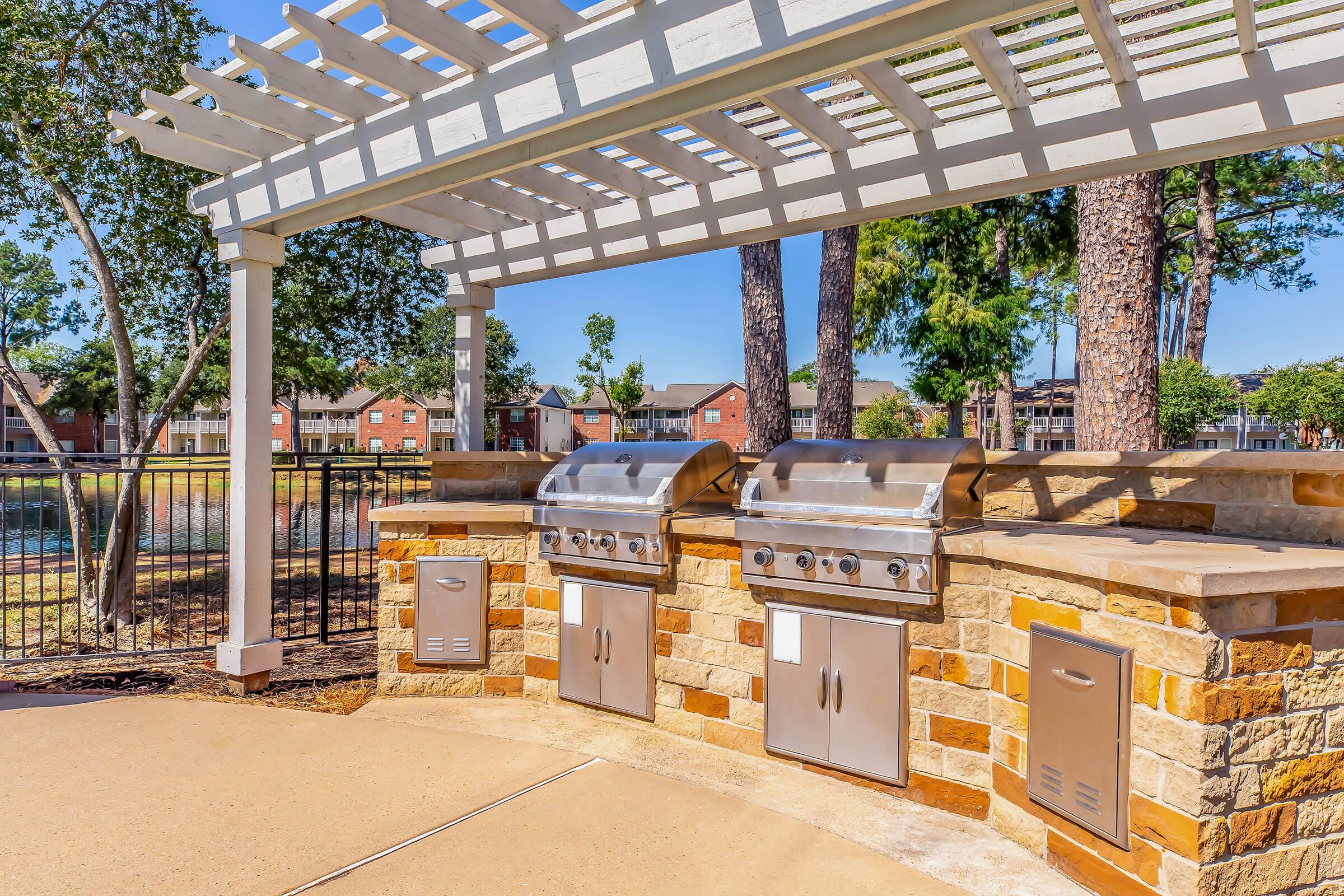 Outdoor communal grilling area featuring two stainless steel grills built into a stone counter, shaded by a pergola. Surrounding trees and a pool are visible in the background, creating a relaxing environment for outdoor cooking and dining.