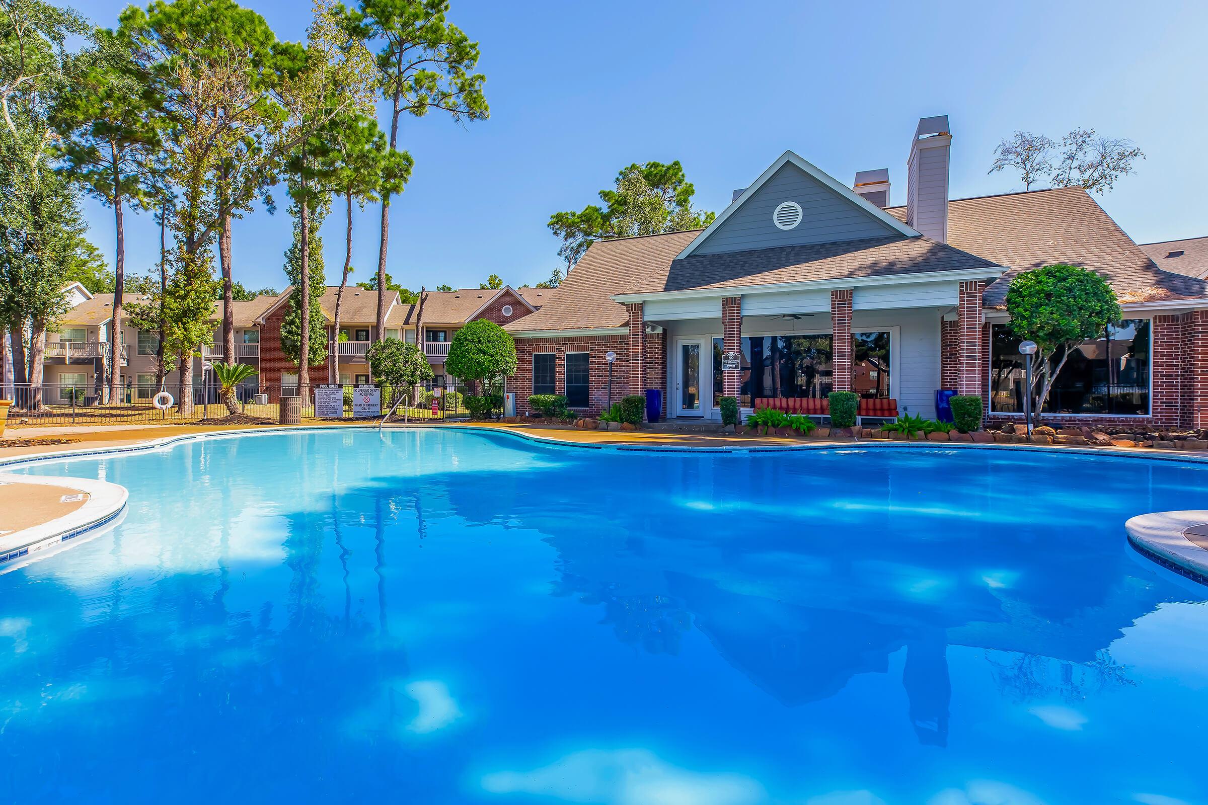 A clear blue swimming pool surrounded by trees and spacious lawn. In the background, there is a large building with a light-colored facade and multiple windows. The scene is bright and sunny, creating a relaxing outdoor atmosphere.
