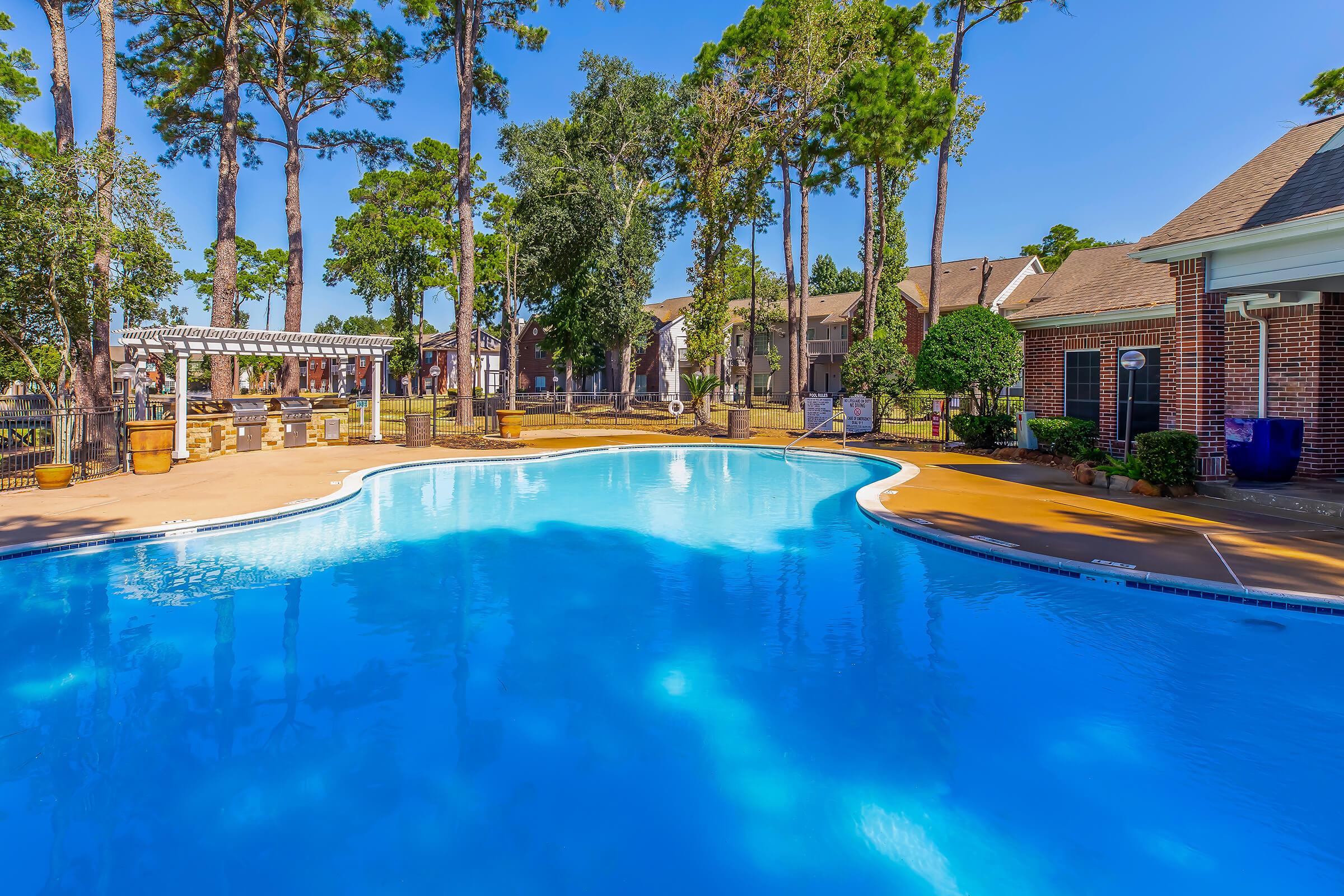 A clear blue swimming pool surrounded by lush greenery and trees, with a sunlit patio area. Nearby, there are residential buildings and a decorative gazebo, creating a relaxing outdoor ambiance.