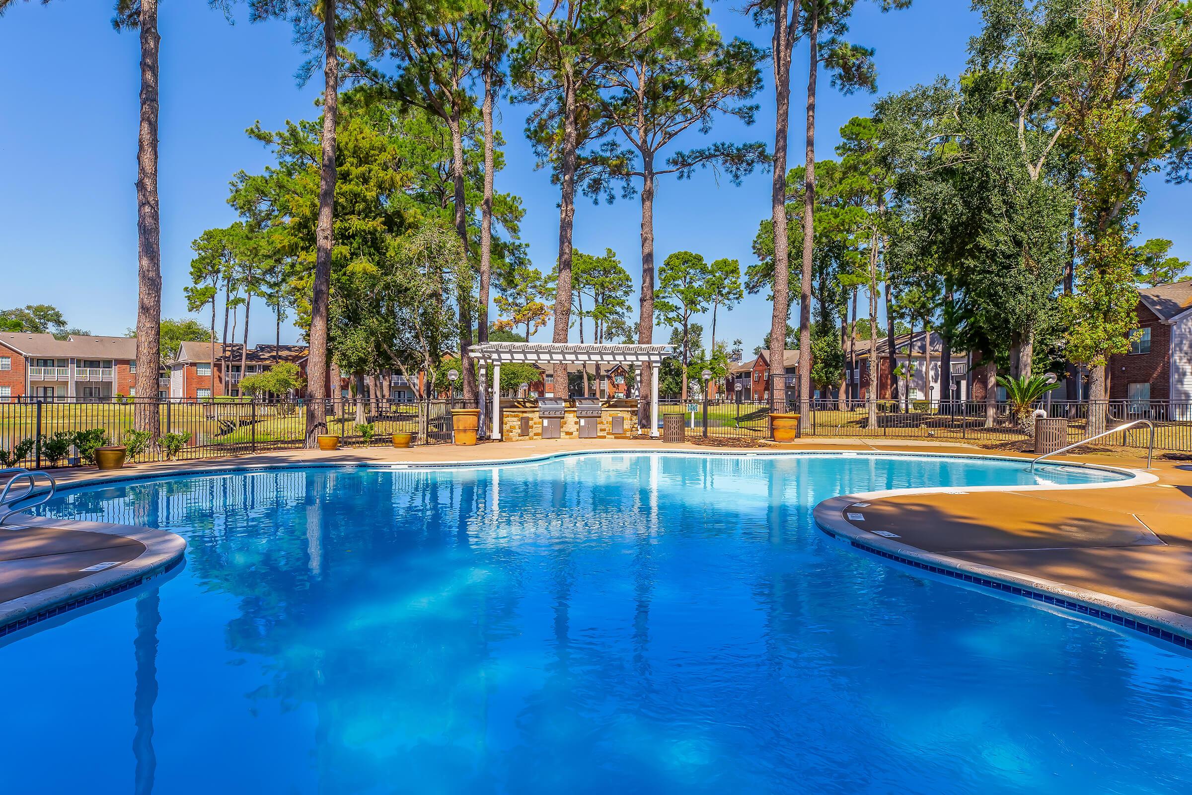 A clear, blue pool surrounded by lush greenery and tall trees, with a shaded sitting area featuring a pergola in the background. The pool area is well-maintained, inviting for relaxation, and offers a serene atmosphere in a residential setting.