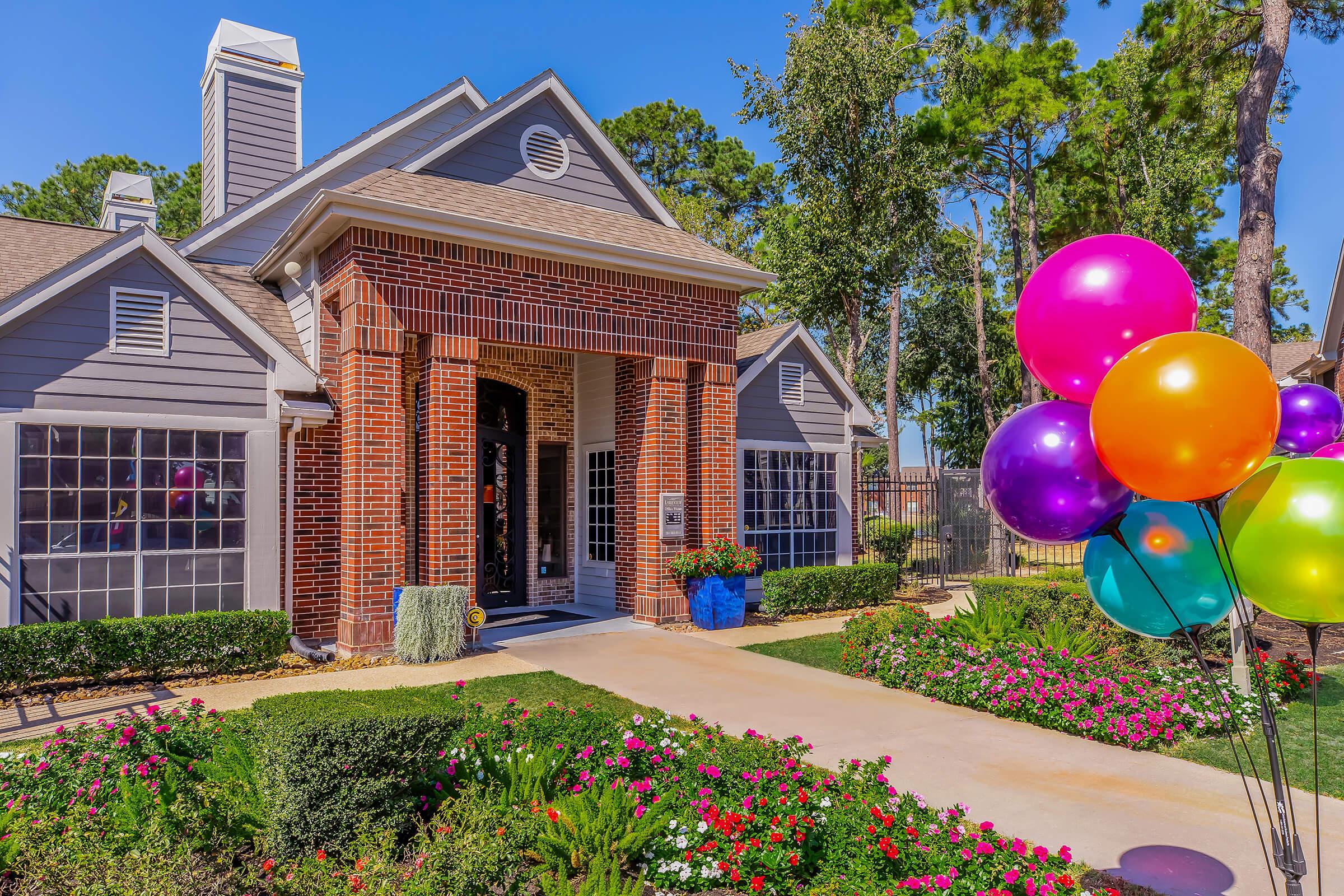 A charming gray house with a red brick entrance surrounded by lush greenery and colorful flowers. A cluster of bright balloons in purple, pink, green, and gold is in the foreground, adding a festive touch to the scene. The sky is clear and blue, enhancing the cheerful atmosphere.