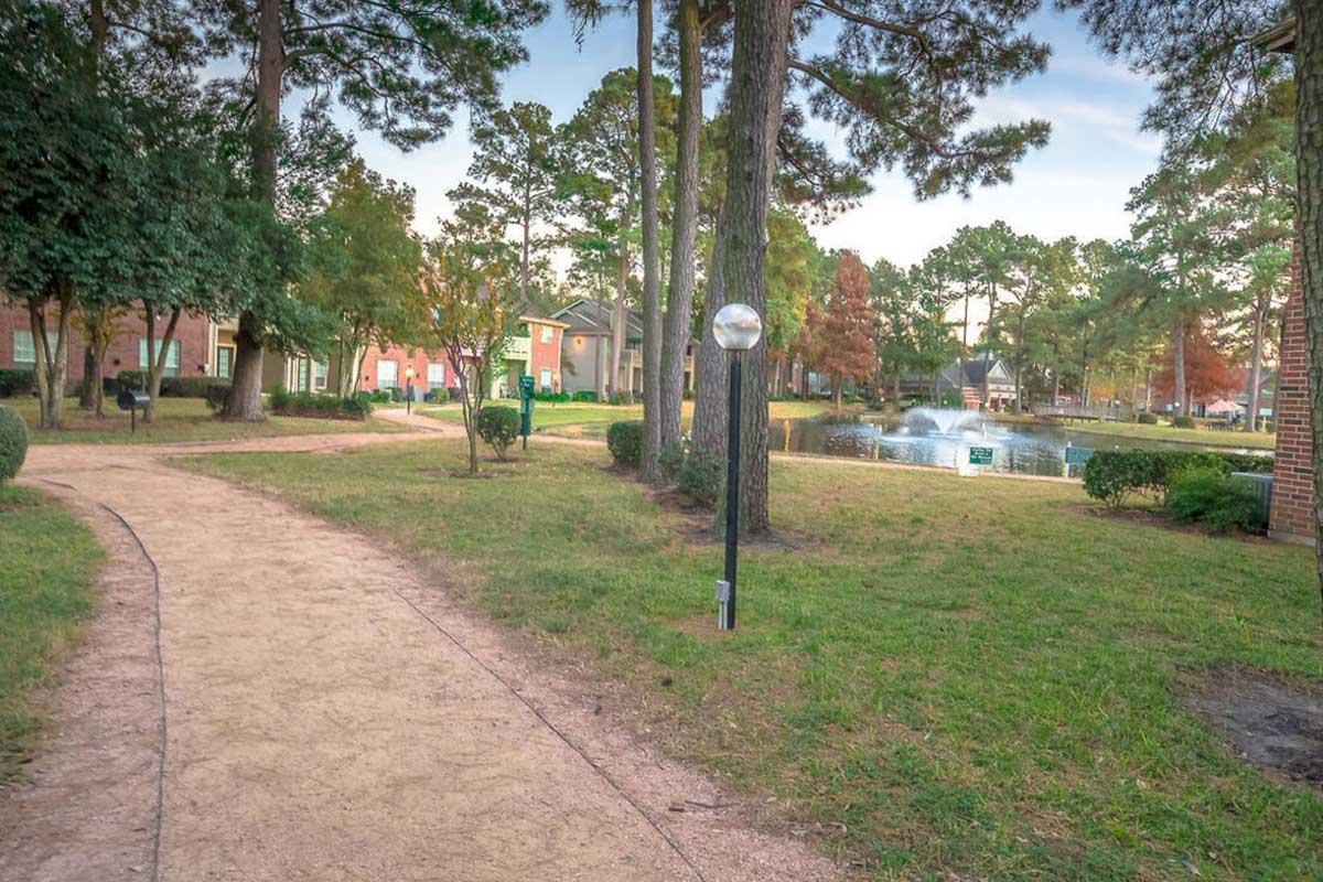 A peaceful park scene featuring a winding path lined with lush greenery, trees, and shrubs. In the background, there are lakeside views with water fountains and residential buildings visible. The atmosphere is tranquil, suggesting a perfect setting for walking or relaxing outdoors.