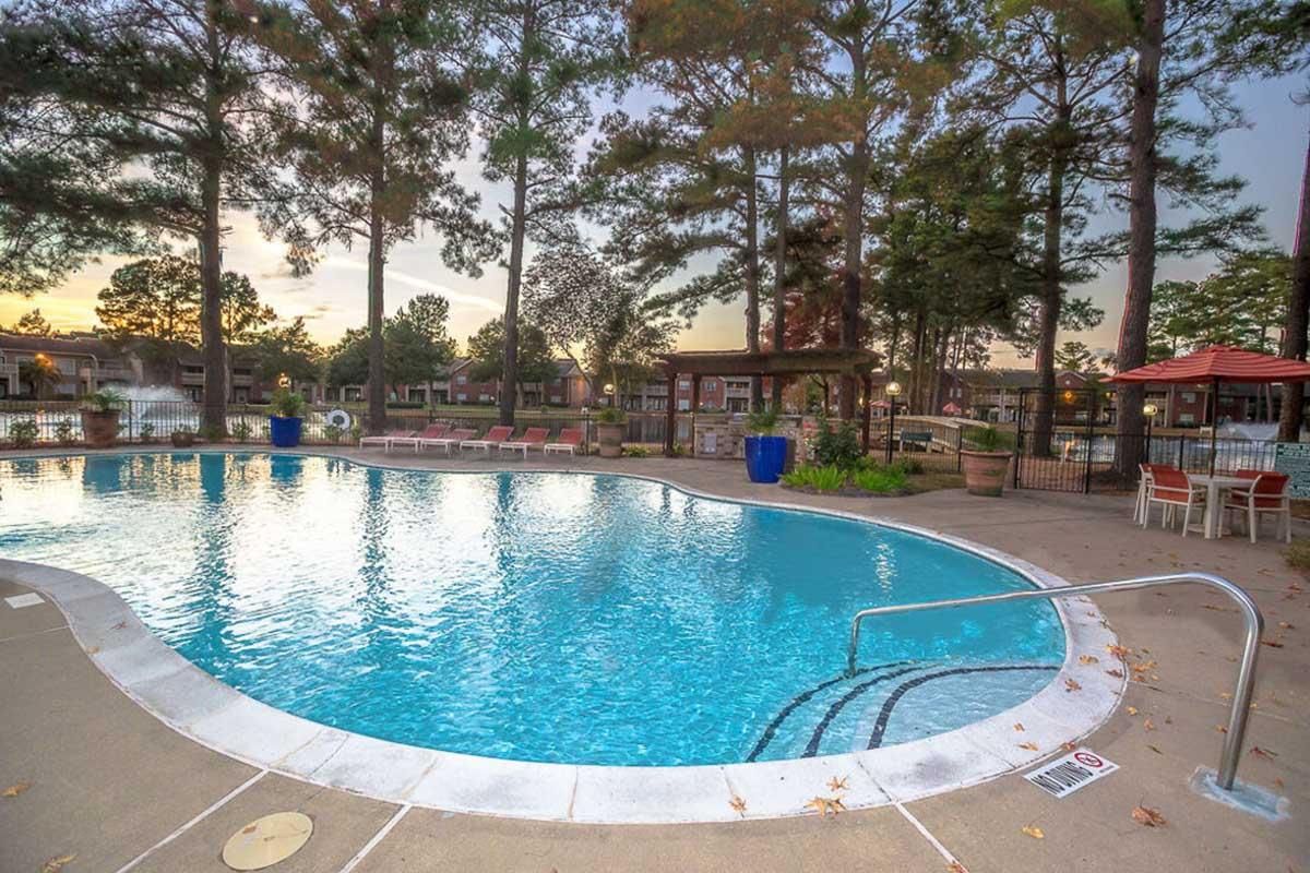 A serene swimming pool surrounded by trees at sunset. The pool features clear blue water and a safe entry step. Comfortable lounge chairs are positioned around the pool area, with shaded seating under umbrellas. The inviting atmosphere is perfect for relaxation and leisure.