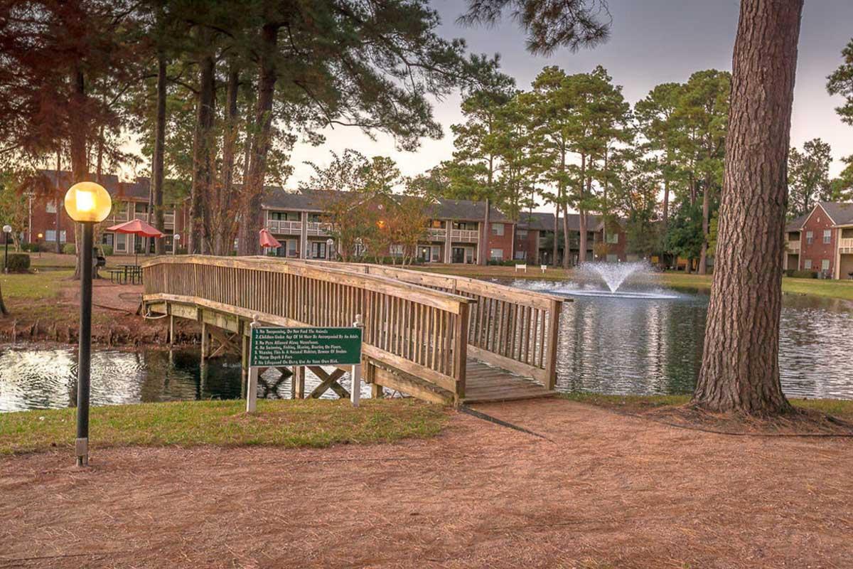 A wooden bridge spans a serene pond, surrounded by trees. A fountain sprays water in the center of the pond. In the background, several apartment buildings are visible, along with a red gazebo. Soft evening light casts a warm glow over the scene, enhancing the peaceful atmosphere.