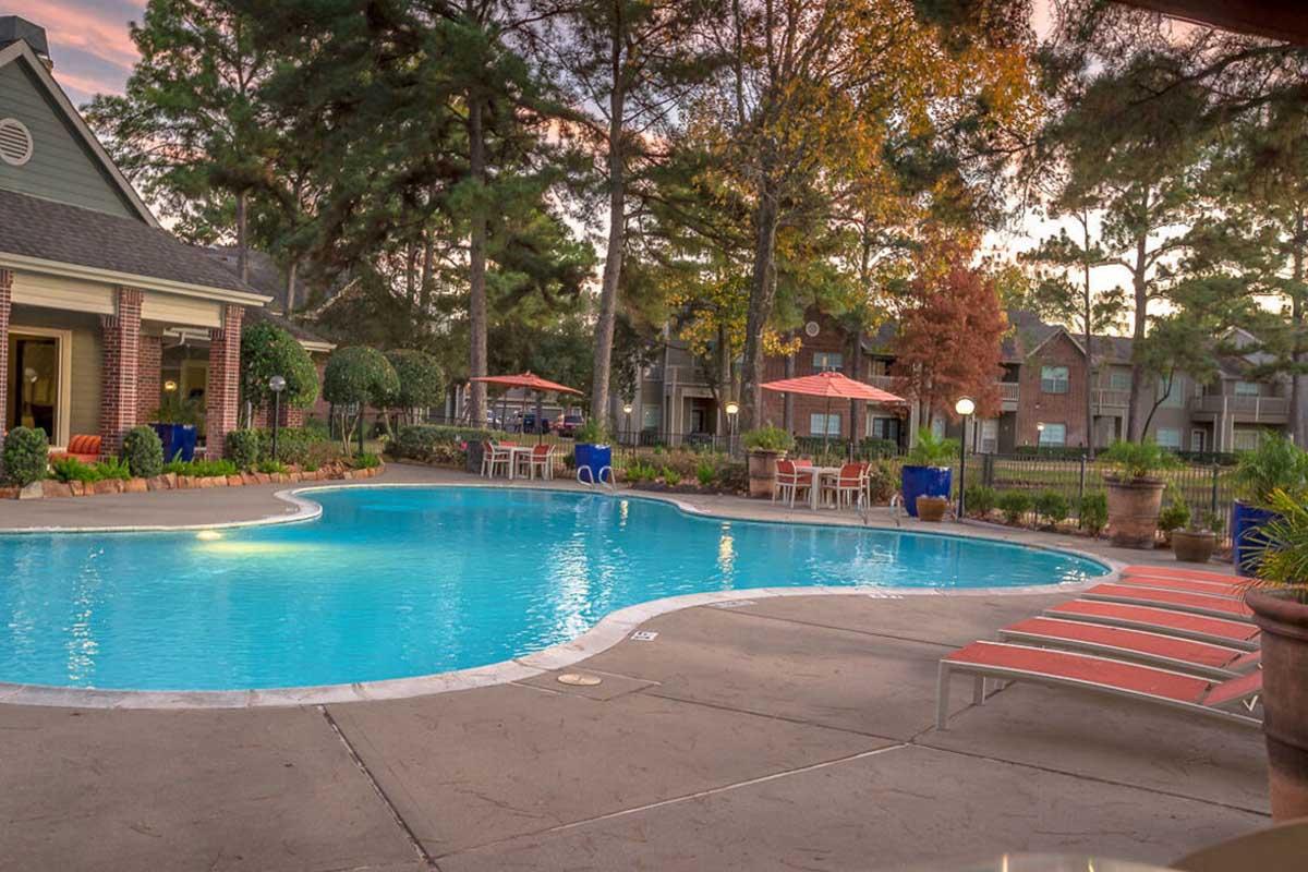 A serene swimming pool surrounded by lush greenery, lounges, and shaded umbrellas. In the background, there are apartment buildings and vibrant trees. The scene is illuminated by the soft glow of sunset, creating a relaxing atmosphere ideal for leisure and relaxation.