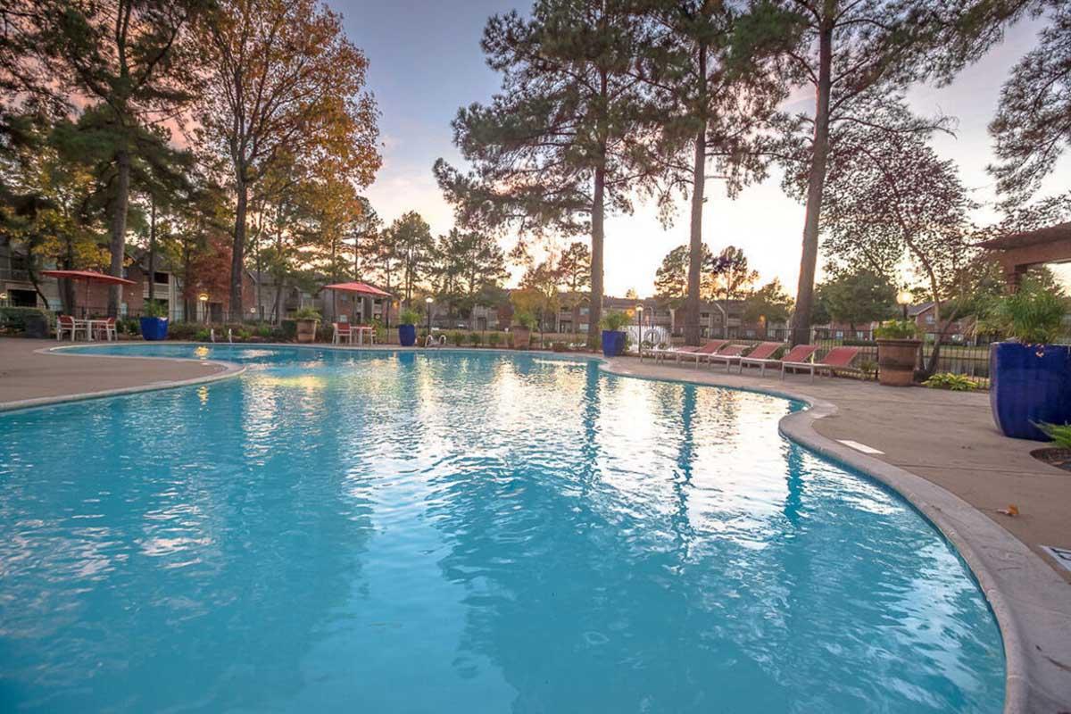 A serene swimming pool surrounded by lush green trees, with lounge chairs neatly arranged nearby. The water glistens in the evening light, reflecting a warm sunset. A relaxing atmosphere is created by the combination of natural scenery and the inviting pool area.
