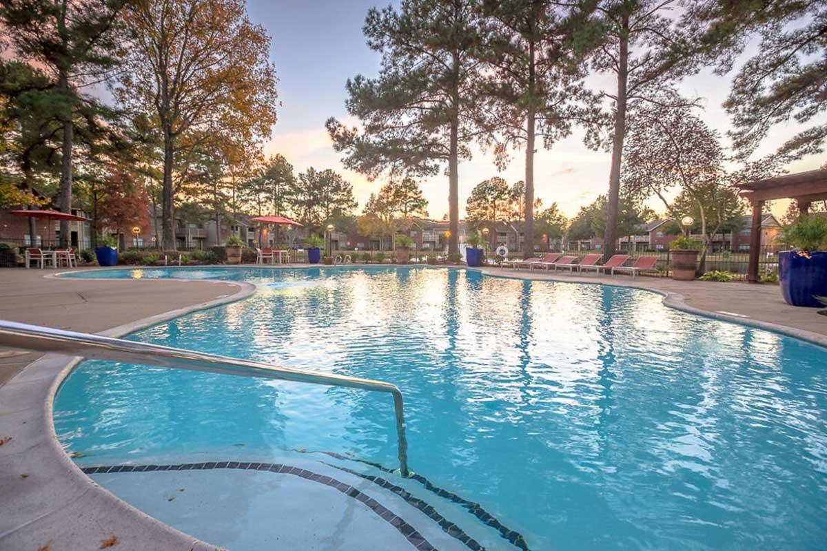 A serene outdoor swimming pool surrounded by tall trees and lounge chairs. The water is clear and reflects the colors of the sunset, creating a tranquil atmosphere. Nearby, there are shaded areas and landscaped gardens, inviting relaxation and leisure.