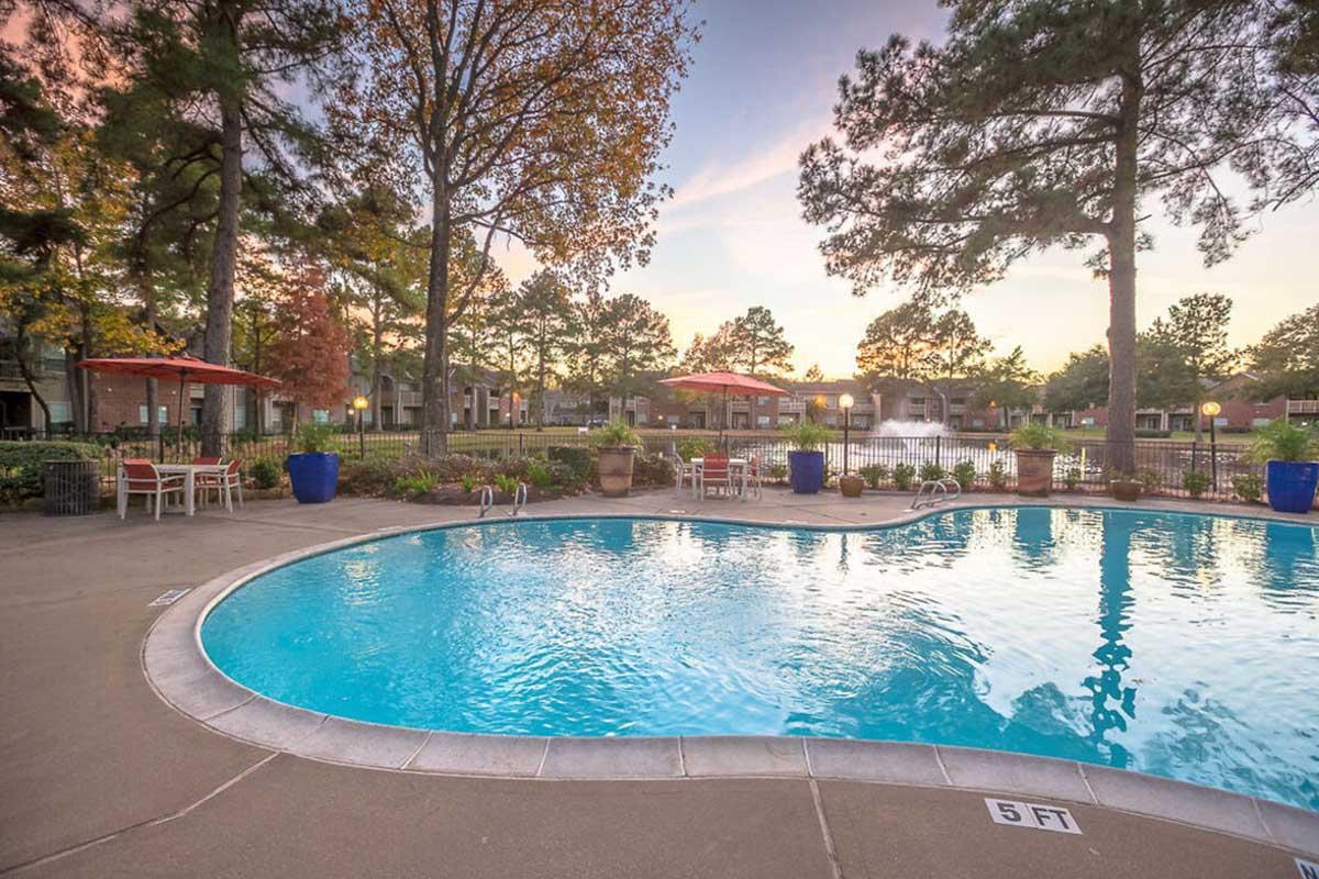 A serene outdoor pool area surrounded by trees and colorful potted plants. The water is clear, reflecting the sky at sunset. There are lounge chairs and tables with umbrellas nearby, and a gated area in the background with additional greenery. The depth of the pool is marked at 5 feet.