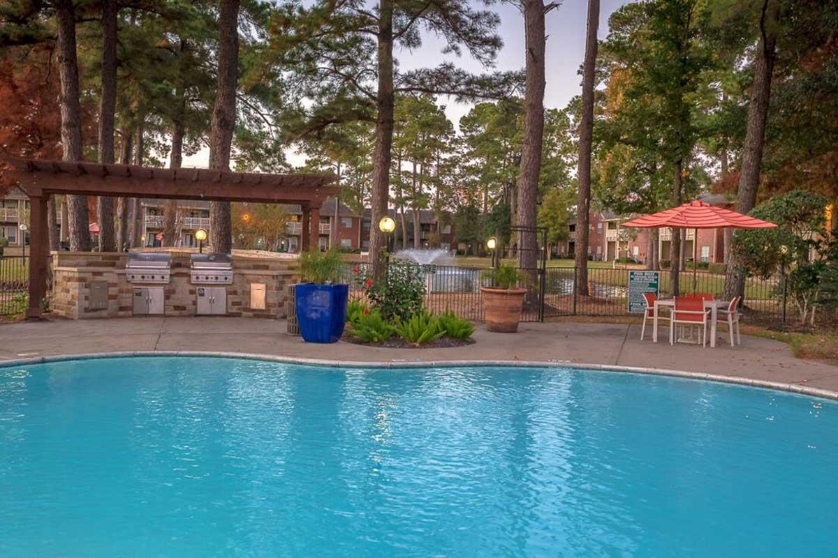 A swimming pool surrounded by trees, featuring a patio area with grills, a blue planter, and an umbrella table. In the background, there are apartment buildings and a fountain, all set in a serene outdoor environment during the evening.