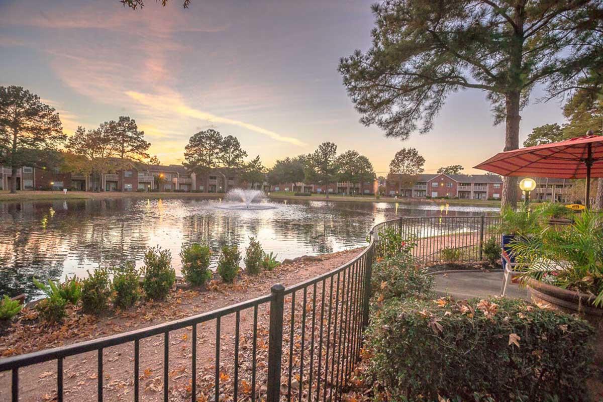 Scenic view of a tranquil pond surrounded by greenery at sunset. A fountain creates gentle ripples in the water, and nearby, a cozy seating area with an umbrella overlooks the serene scene. A wrought iron fence lines the pond, enhancing the peaceful atmosphere.