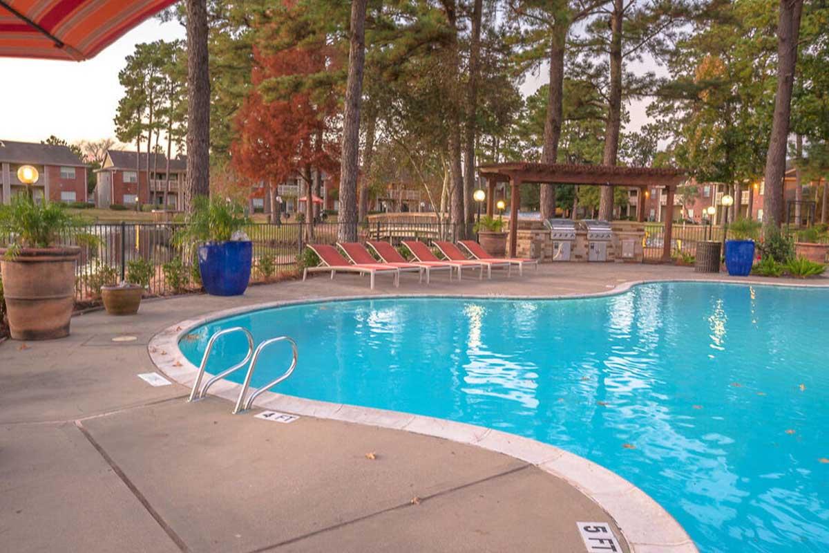 A tranquil swimming pool surrounded by green trees and lounge chairs. The pool features clear blue water and a shallow area marked with depth indicators. Nearby, there are barbecue grills under a wooden gazebo, creating a relaxing outdoor atmosphere.