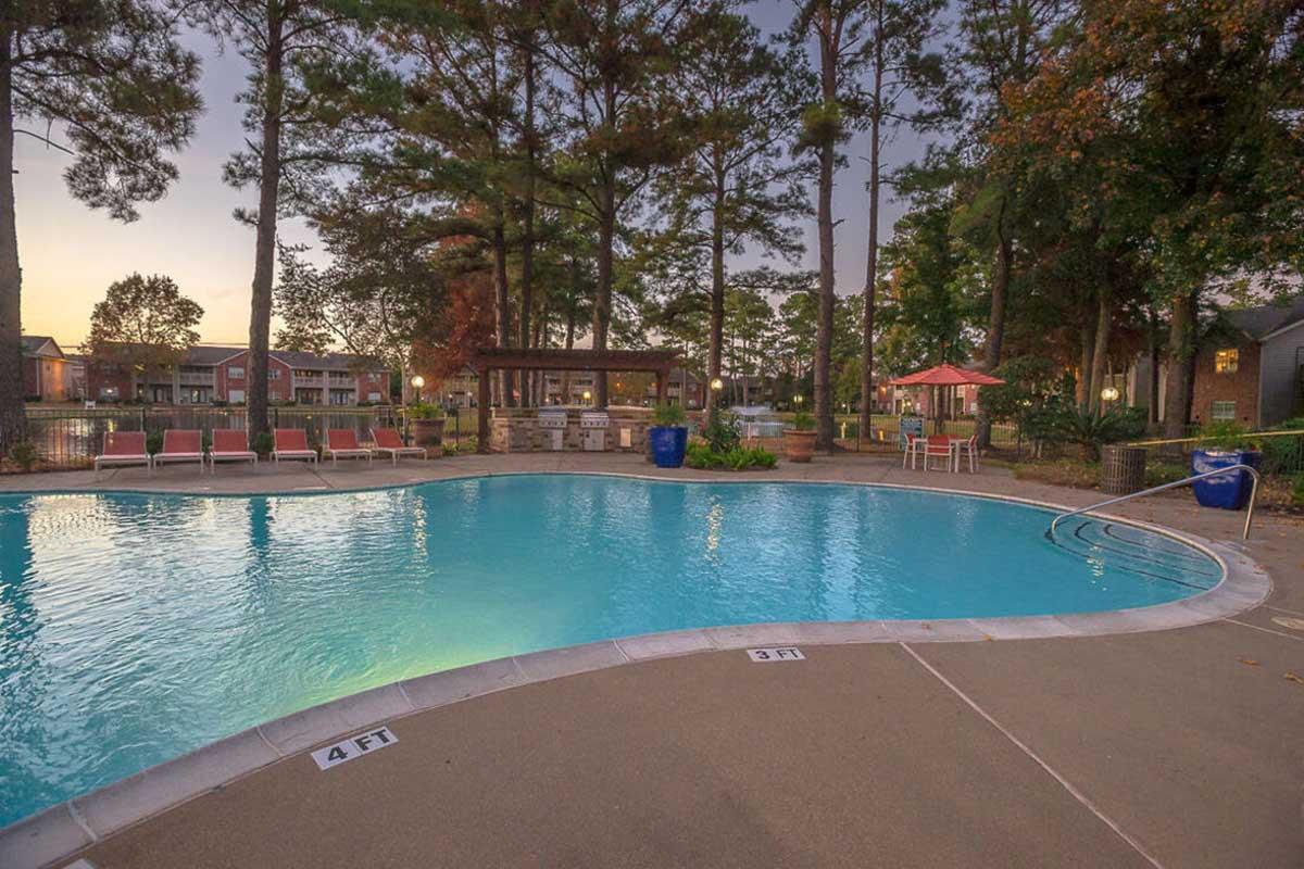 A serene swimming pool with clear blue water surrounded by a concrete deck. Lounge chairs line one side, and a shaded gazebo is visible in the background among tall trees. The scene is set in the evening with warm lighting, creating a relaxing atmosphere in a residential area.