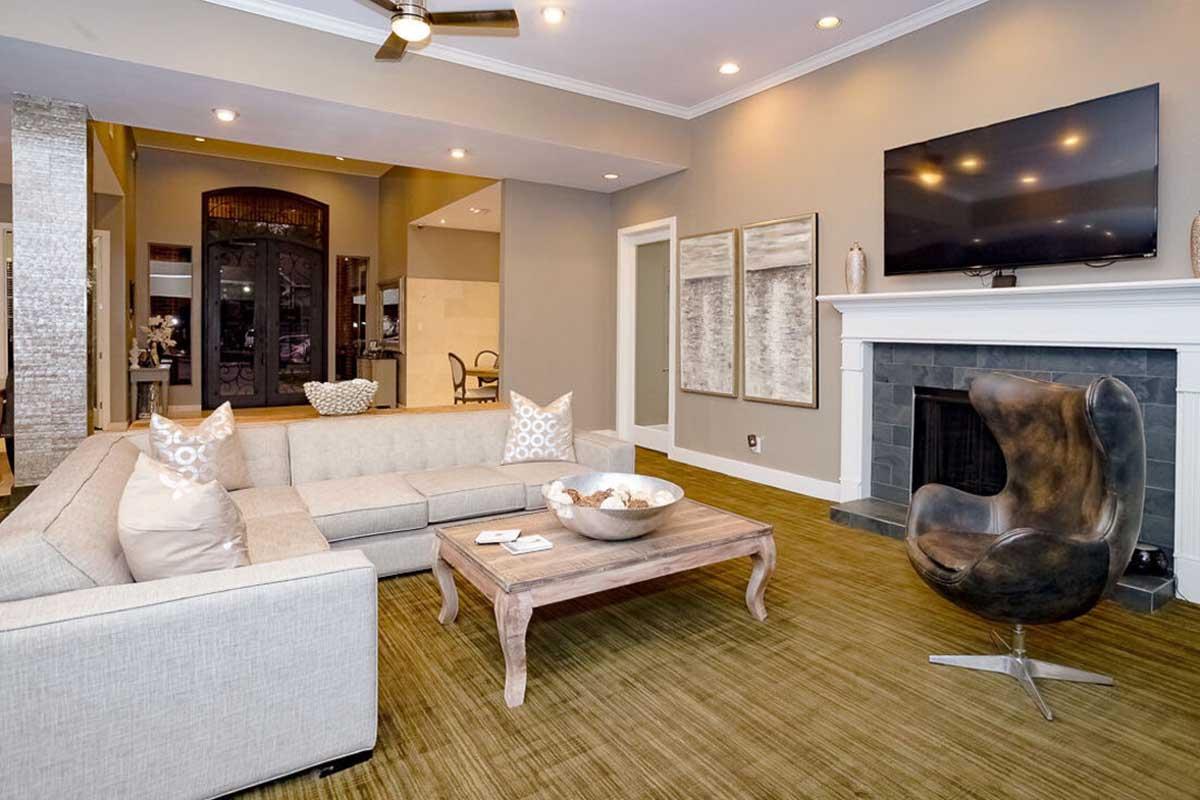 Modern living room featuring a light gray sectional sofa with decorative pillows, a wooden coffee table with a bowl of fruit, and a stylish black leather chair. The space has neutral-colored walls, a fireplace, and a large flat-screen TV mounted above a screened fireplace, creating a cozy and inviting atmosphere.