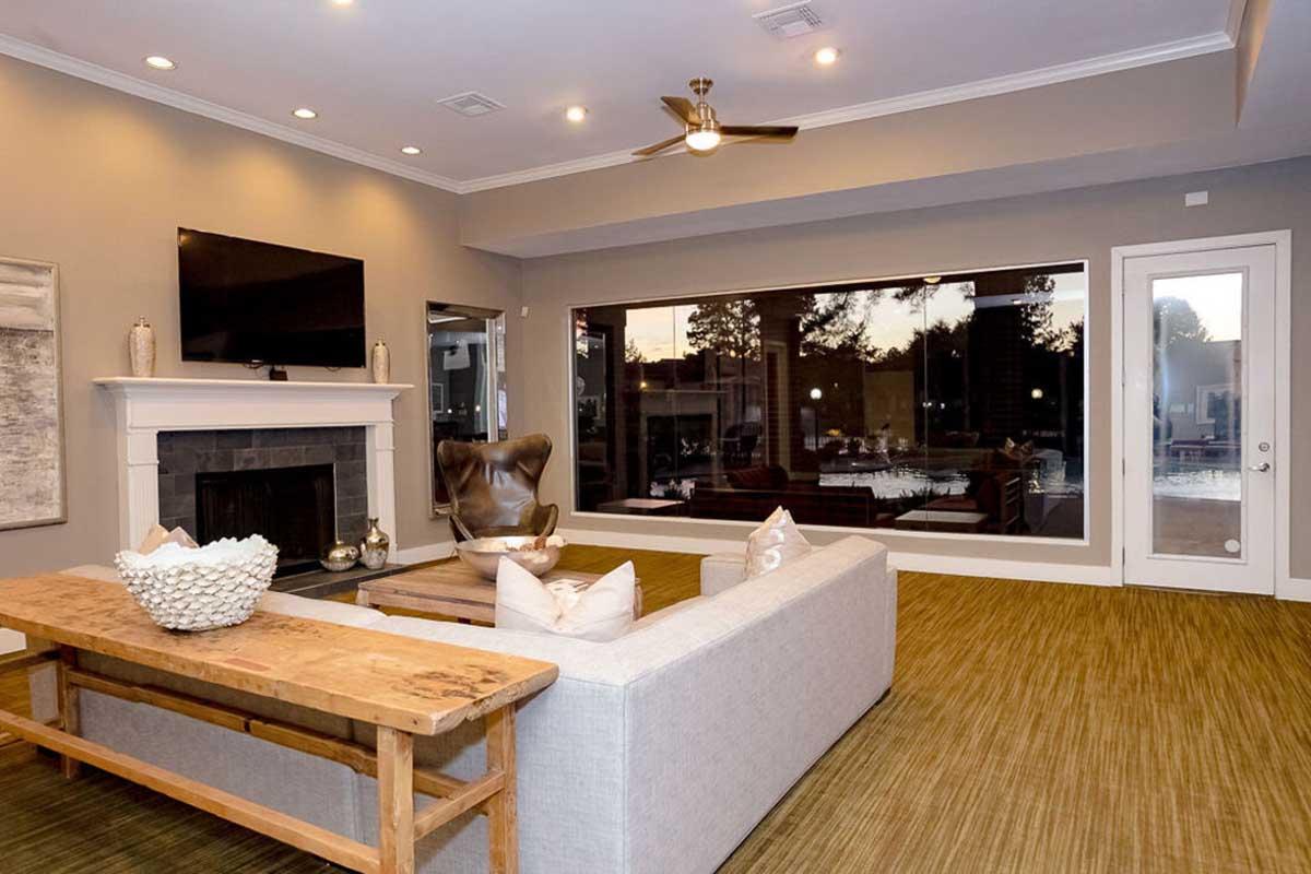 A modern living room featuring a light-colored sectional sofa, a rustic wooden coffee table, and a flat-screen TV mounted above a fireplace. Large windows provide natural light and a view of an outdoor space with a pool. The flooring is a textured green carpet, enhancing the contemporary decor.