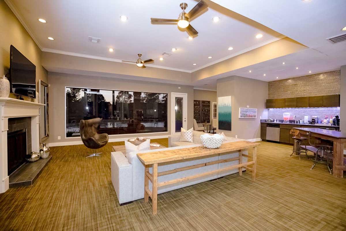 A modern, spacious living area featuring a large window with outdoor views, a comfortable sofa, a wooden dining table, stylish chairs, a fireplace, and a kitchenette with bar seating. Ceiling fans add to the contemporary decor, which includes a mix of neutral and warm tones.
