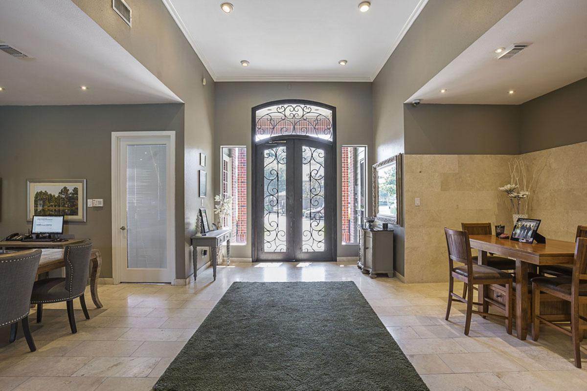 A spacious entryway featuring a large decorative front door with wrought-iron accents. To the left, there is a small office area with a desk. The right side includes a dining table with chairs. The floor is tiled with light-colored stone, and a green area rug adds a touch of warmth. Natural light streams in through the door.