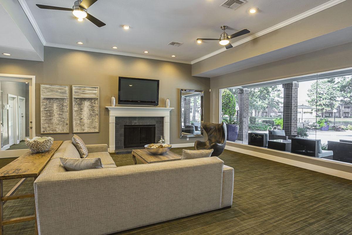 Modern living room featuring a gray sofa, a wooden coffee table, and a fireplace. The walls are painted in neutral tones, with decorative artwork on display. Large windows provide a view of an outdoor space, and ceiling fans add to the airy atmosphere. Elegant decor creates a welcoming environment.