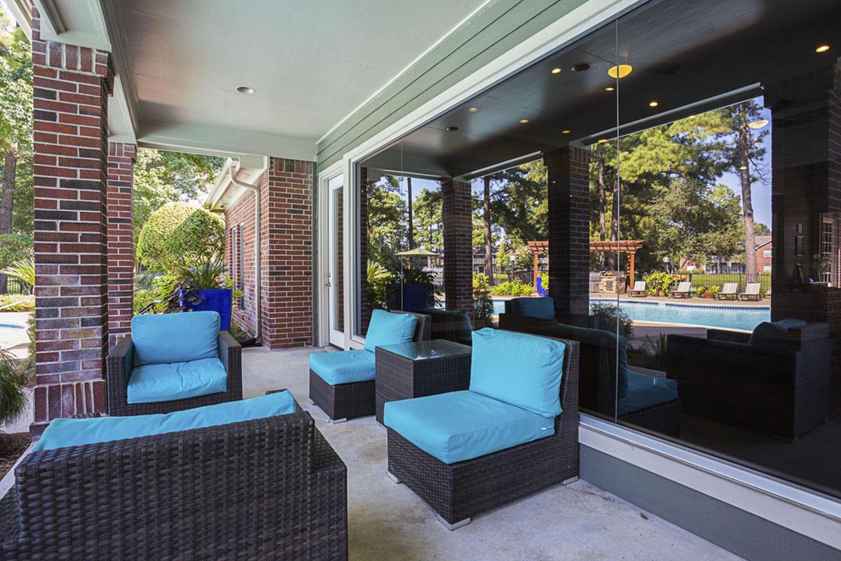 A cozy outdoor seating area featuring dark wicker chairs with bright turquoise cushions, set against a backdrop of lush greenery and a pool. The space is partially shaded, with large glass doors reflecting the serene environment.